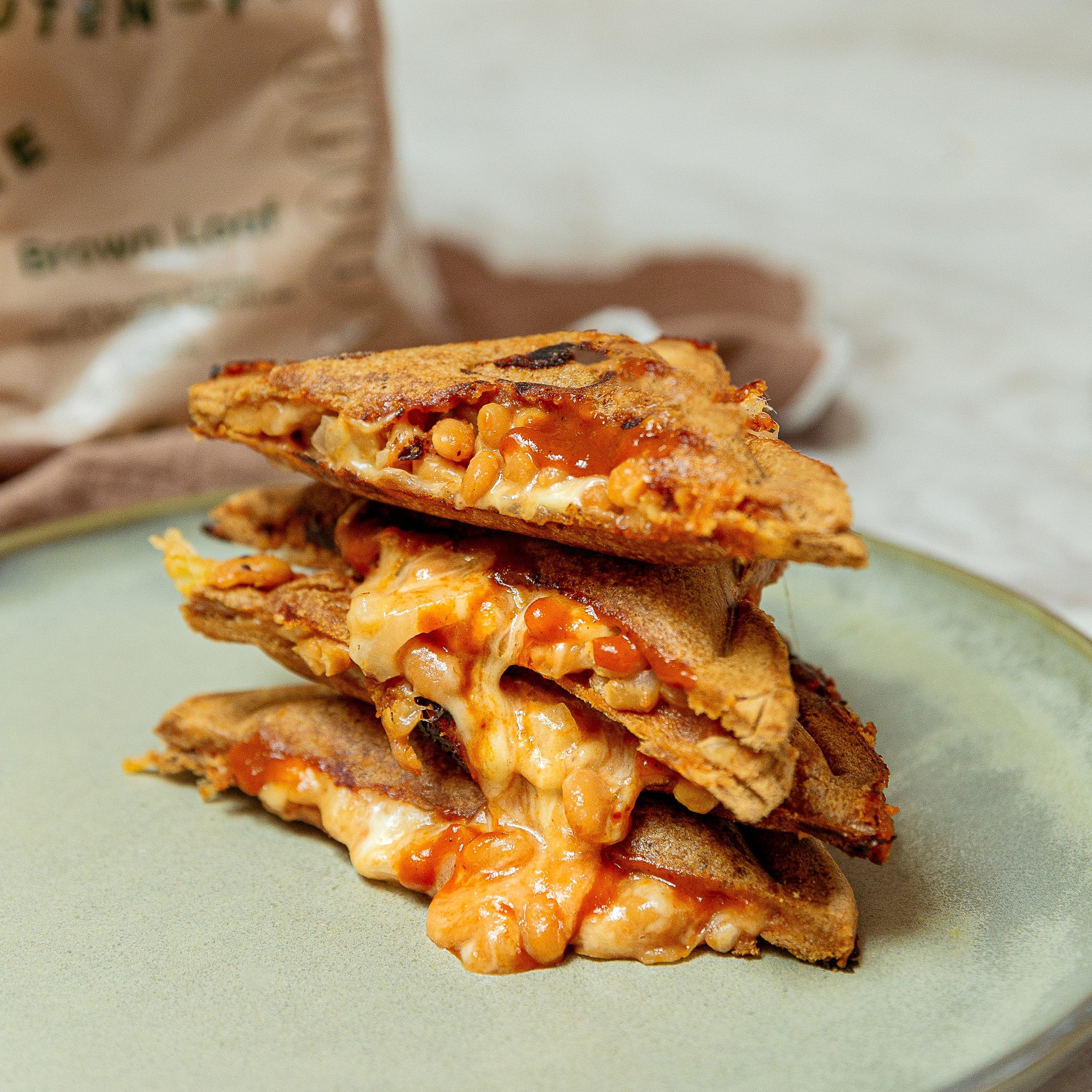 Four triangular pieces of a toasted sandwich stack on a pale green plate, with melted cheese. A brown bread bag labeled gluten-free brown loaf is blurred in the background on a light surface.