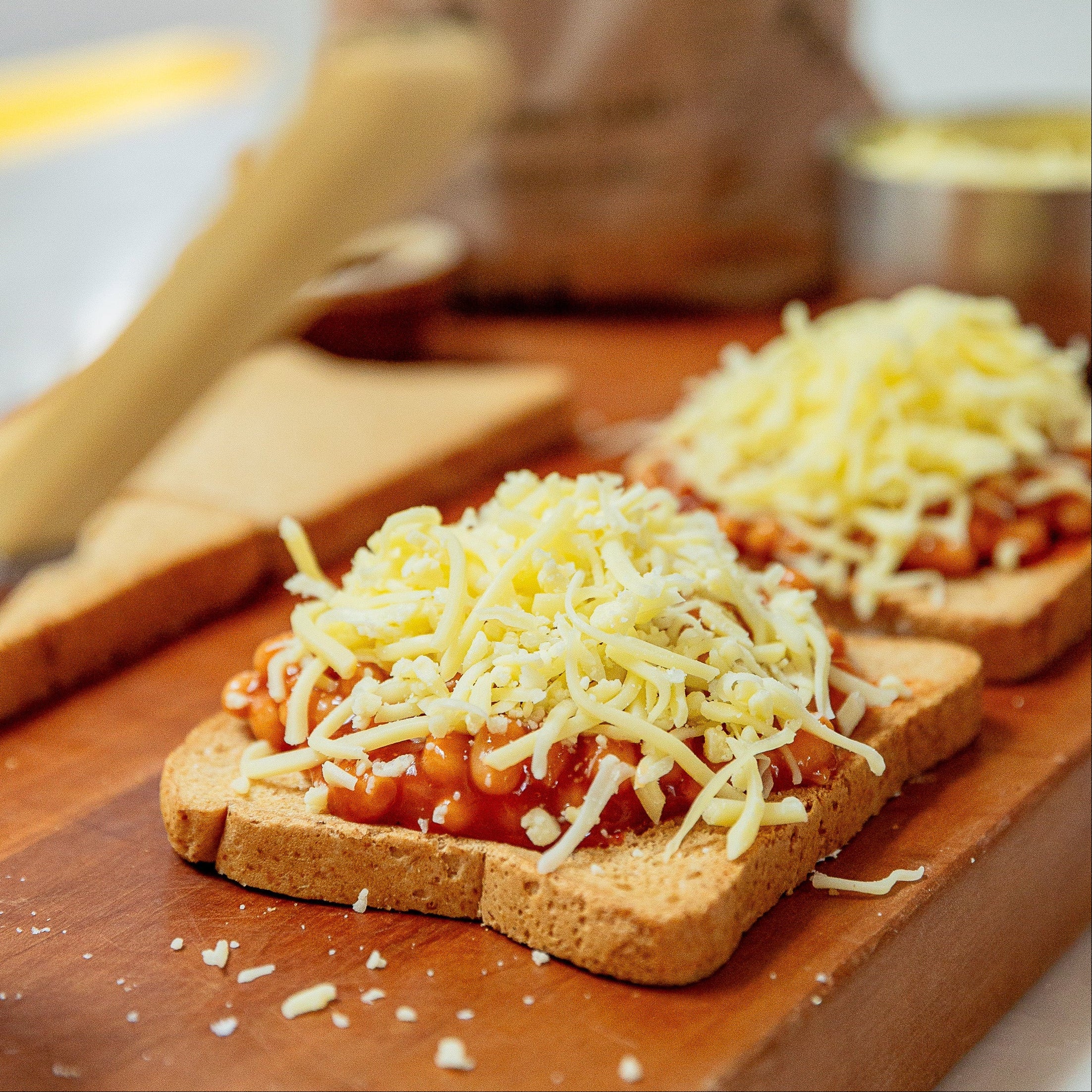 Two slices of toast topped with baked beans and shredded cheese on a wooden cutting board, with a blurred bag of bread in the background.