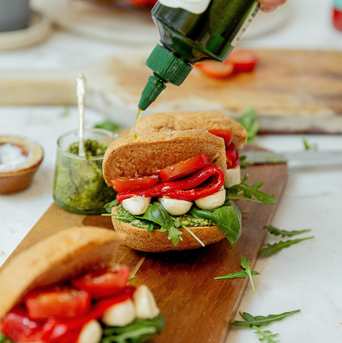 A hand squeezes olive oil from a green bottle onto a Caprese sandwich on a wooden board, with another sandwich and small bowls of ingredients in a bright kitchen setting.