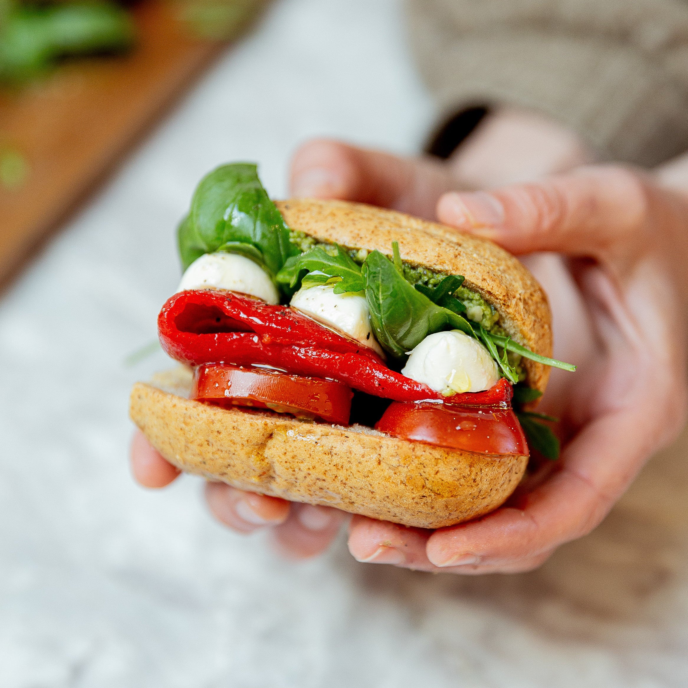 A close-up of hands holding a small caprese sandwich with a brown Allergywise bun, fresh basil leaves, mozzarella balls, and sliced red pepper on a light marble surface.