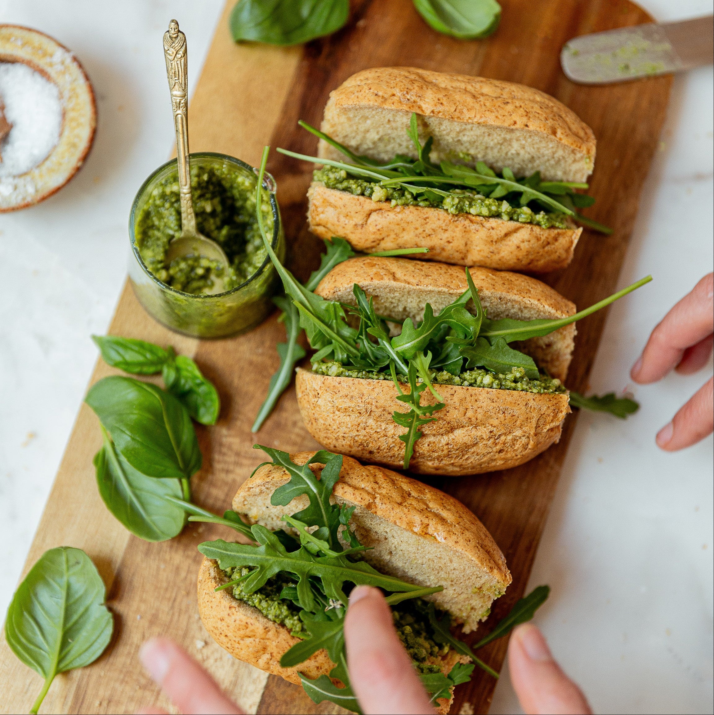 Three arugula and pesto rolls on a wooden board, surrounded by fresh basil leaves, a small jar of green pesto, and hands adjusting the top sandwich.