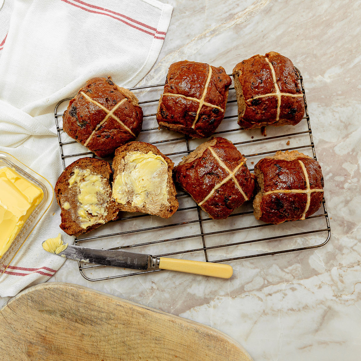 Six golden-brown hot cross buns on a wire cooling rack, with one bun cut open to show buttered interior. A glass dish of butter, a knife, a white cloth, and a wooden board are arranged on a light marble surface.