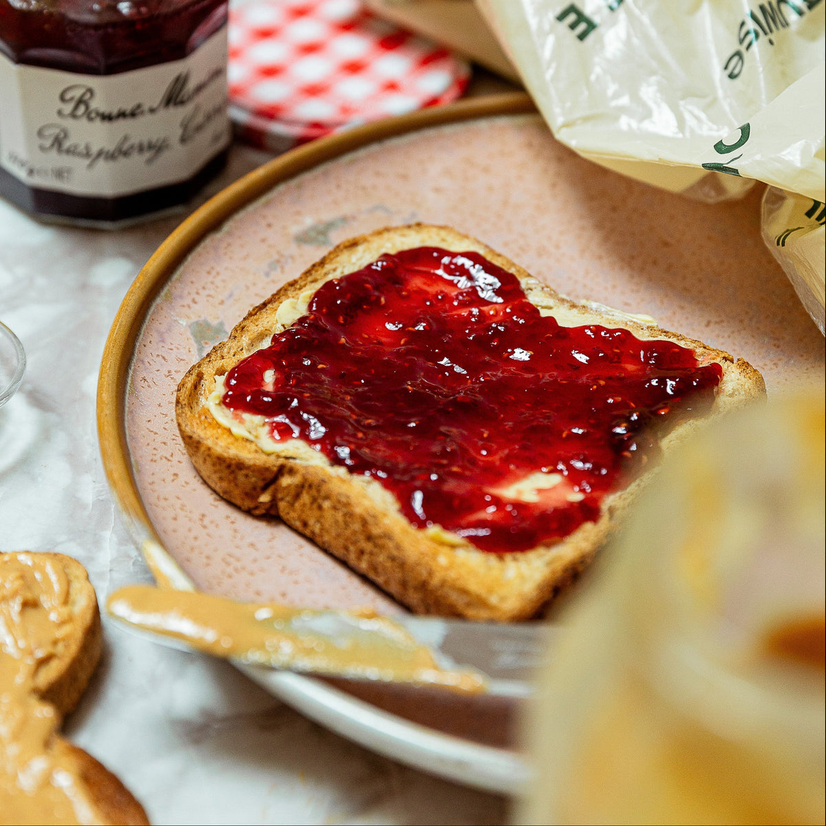 A close-up lifestyle shot of a slice of toast spread with dark raspberry jam on a beige plate, with a jar of raspberry jam, a knife, and a partially unwrapped butter wrapper in a warm kitchen setting. With a white and green Allergywise bread back in the bac.