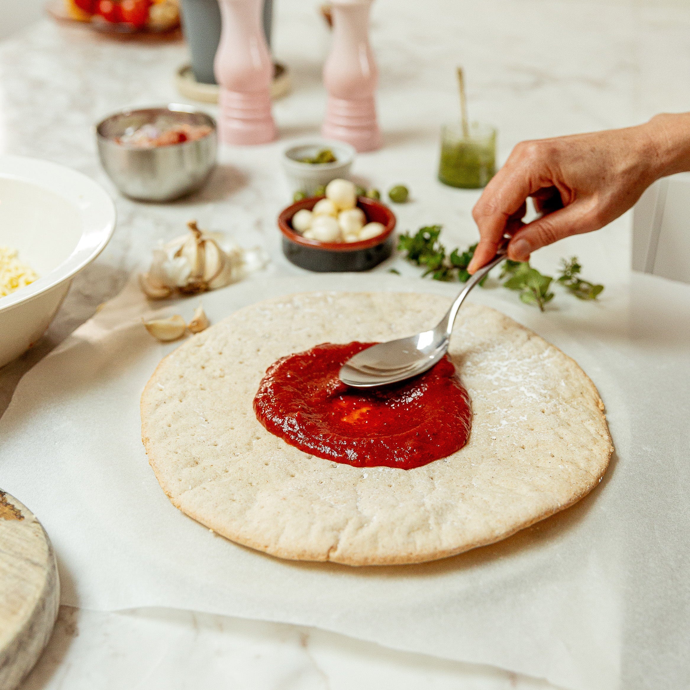 A person's hand uses a spoon to spread red tomato sauce onto a round flatbread pizza crust, set on a white marble countertop with various cooking ingredients and utensils in the background.