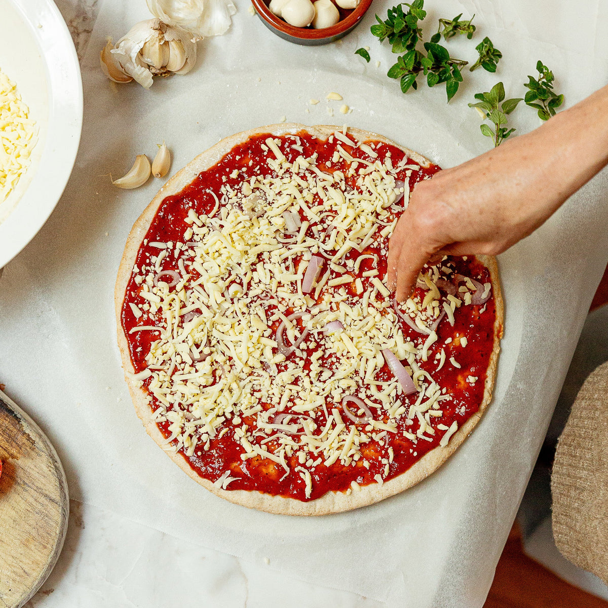 An overhead view of a pizza being topped with shredded cheese and red sauce on a marble countertop, with hands adding ingredients and surrounding food items like garlic, olives, parsley, and pasta visible in the scene.