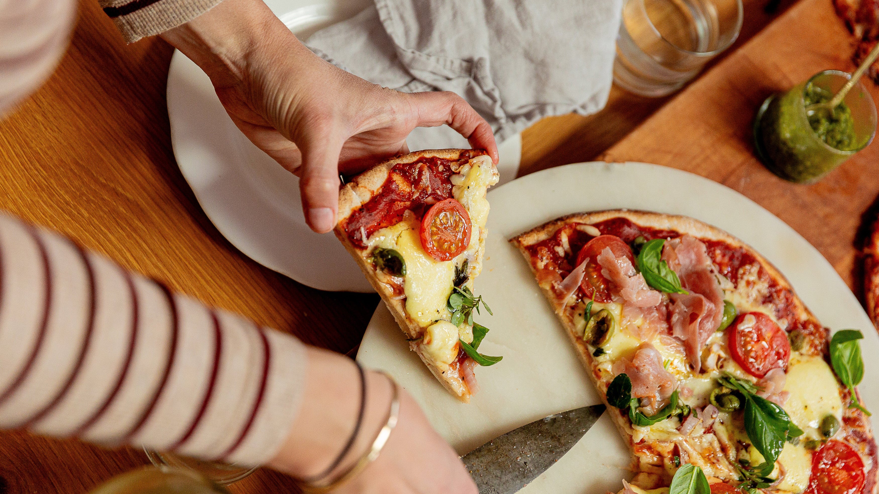 A close-up lifestyle shot of a person holding a slice of pizza with tomato, mozzarella, and basil on a wooden dining table next to a larger pizza on a stone plate.