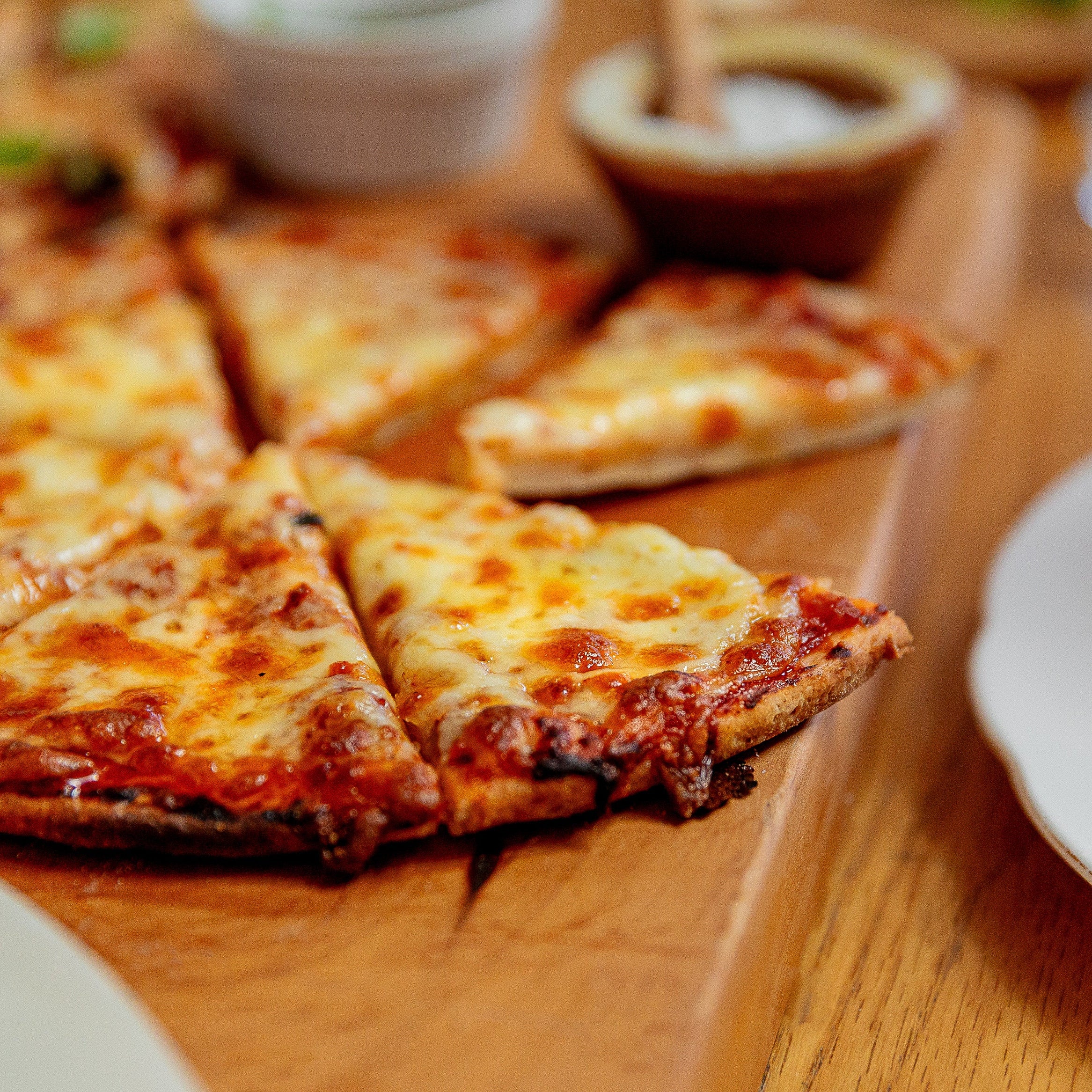 A close-up of a freshly baked cheese pizza on a wooden board, with several slices cut and one slice leaning against the crust. Small dipping sauce bowls and white plates are visible on a wooden table.