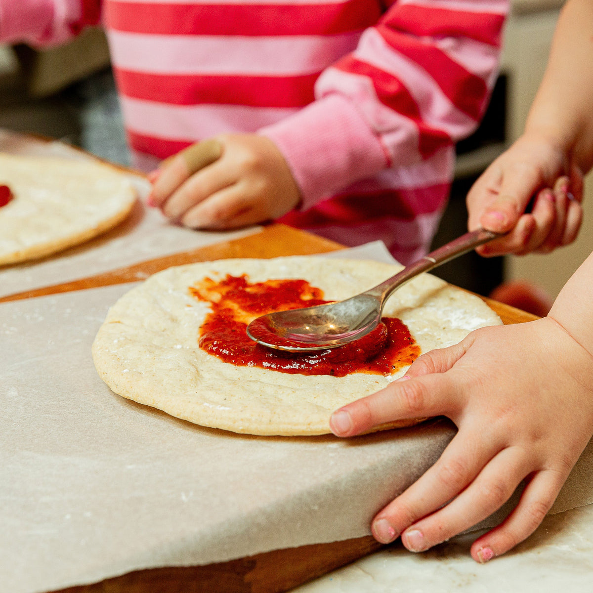 Kids preparing pizza dough by spreading red tomato sauce with spoons on round flour-dusted dough circles over parchment paper.