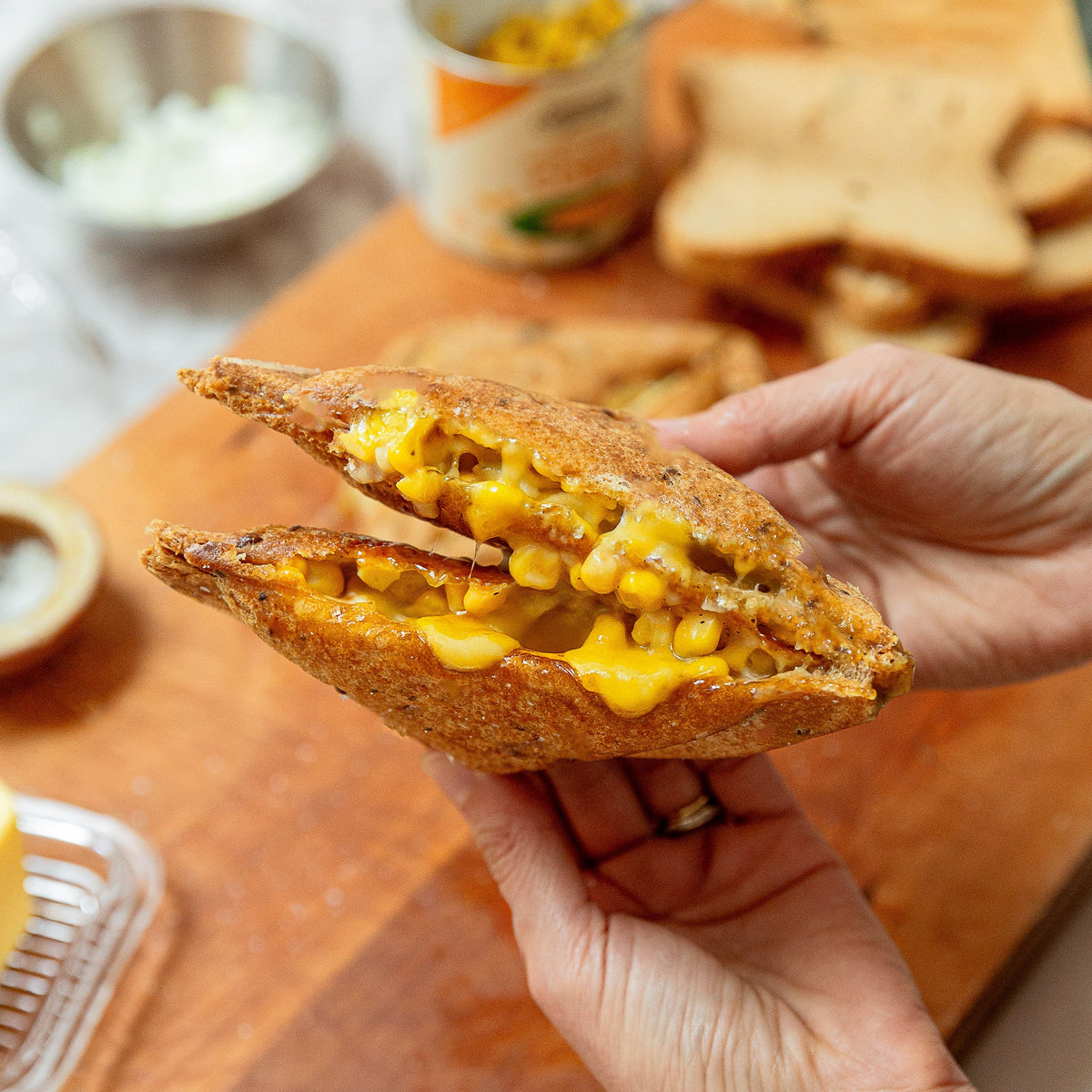 A person holds a toasted sandwich filled with melted cheese and corn kernels over a wooden cutting board, with bread, a can of corn, and condiments in the background.