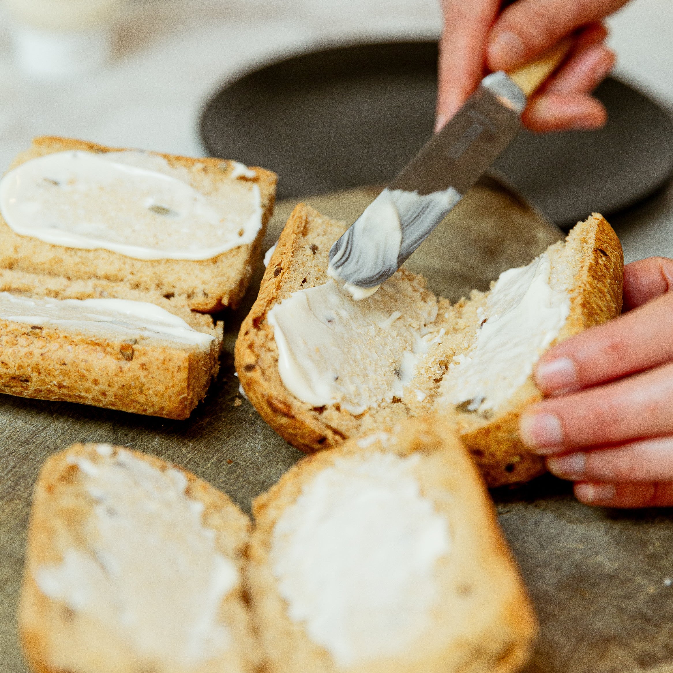A close-up lifestyle shot of a person spreading butter onto a sliced seeded roll with a knife, with other bagels also spread on a wooden board.