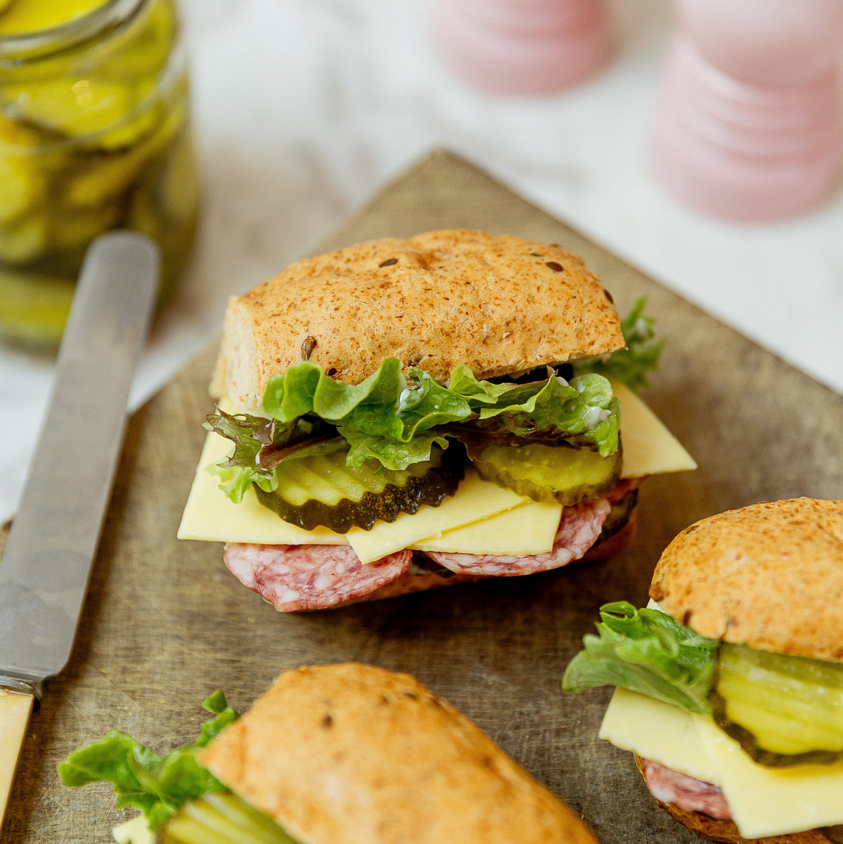 Seeded buns filled with salami, lettuce, pickles and cheese on a wooden chopping board. Pink salt and pepper grinders in the back with a jar of pickles and a butter knife.