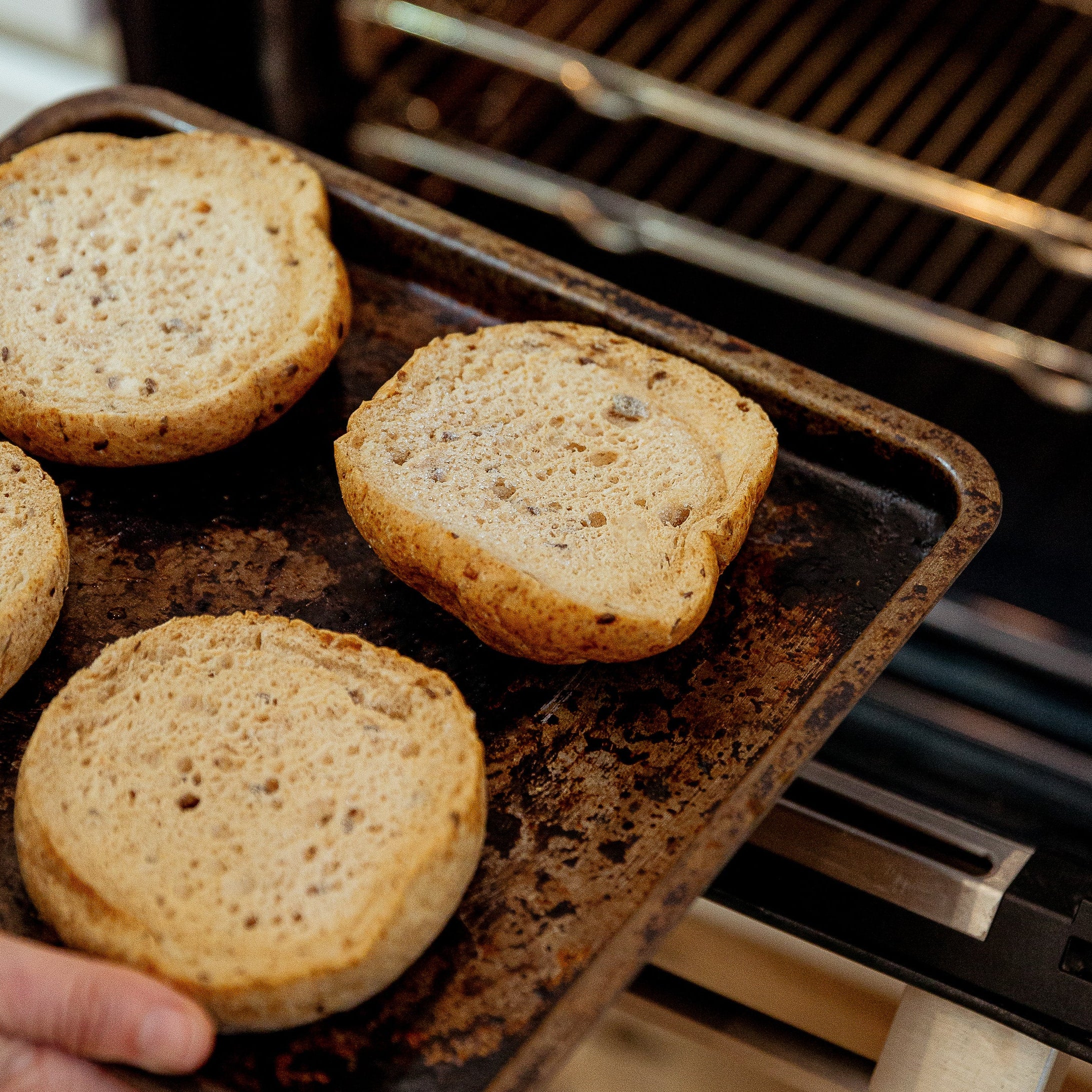 A hand holds a dark baking tray with four toasted bun slices in front of an open oven interior.