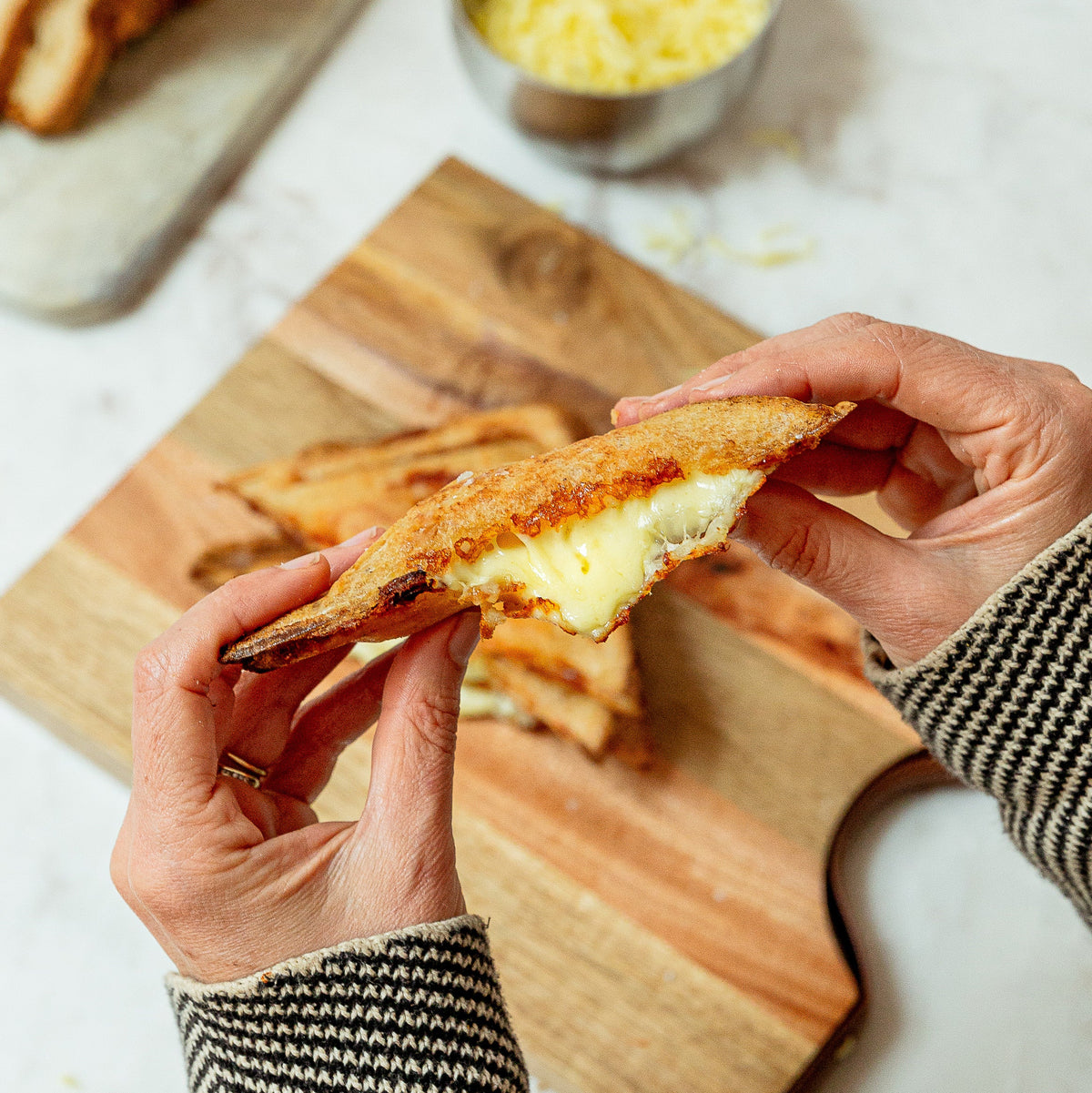 A person's hands holding a golden-brown toasted sandwich on a wooden cutting board, with sliced bread and a small bowl of shredded cheese in the background.