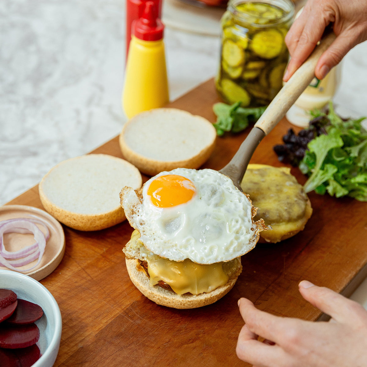 A close-up of a prepared Kiwi burger on a wooden cutting board, featuring a sunny-side-up egg on top of melted cheese and a beef patty, with hands adding the top bun. Condiments, sliced beets, and fresh greens are visible in the background.