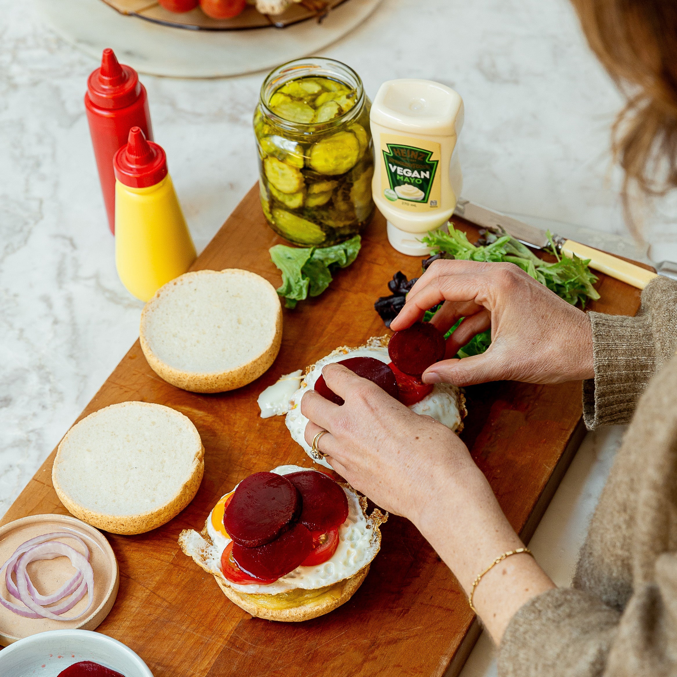 A person preparing a gourmet Kiwi burger on a wooden cutting board, adding sliced red beets and a fried egg to a brioche bun. The scene is styled with condiments, pickles, and fresh ingredients on a white marble countertop.