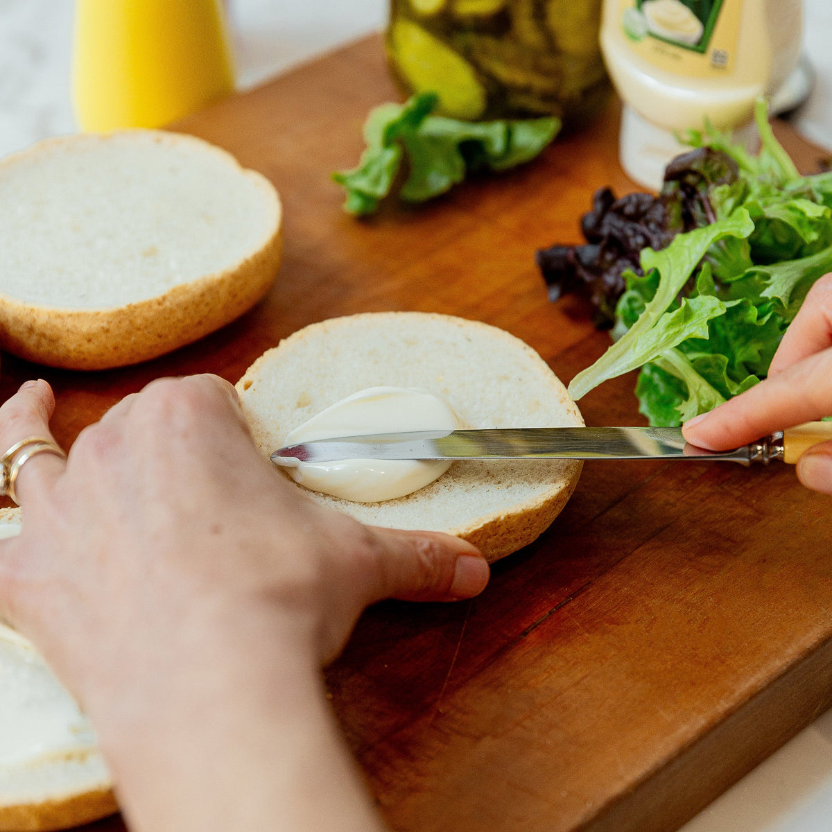 A close-up lifestyle shot of a person spreading vegan mayonnaise onto a burger bun on a wooden cutting board, with Heinz vegan mayonnaise and other ingredients visible in the background.