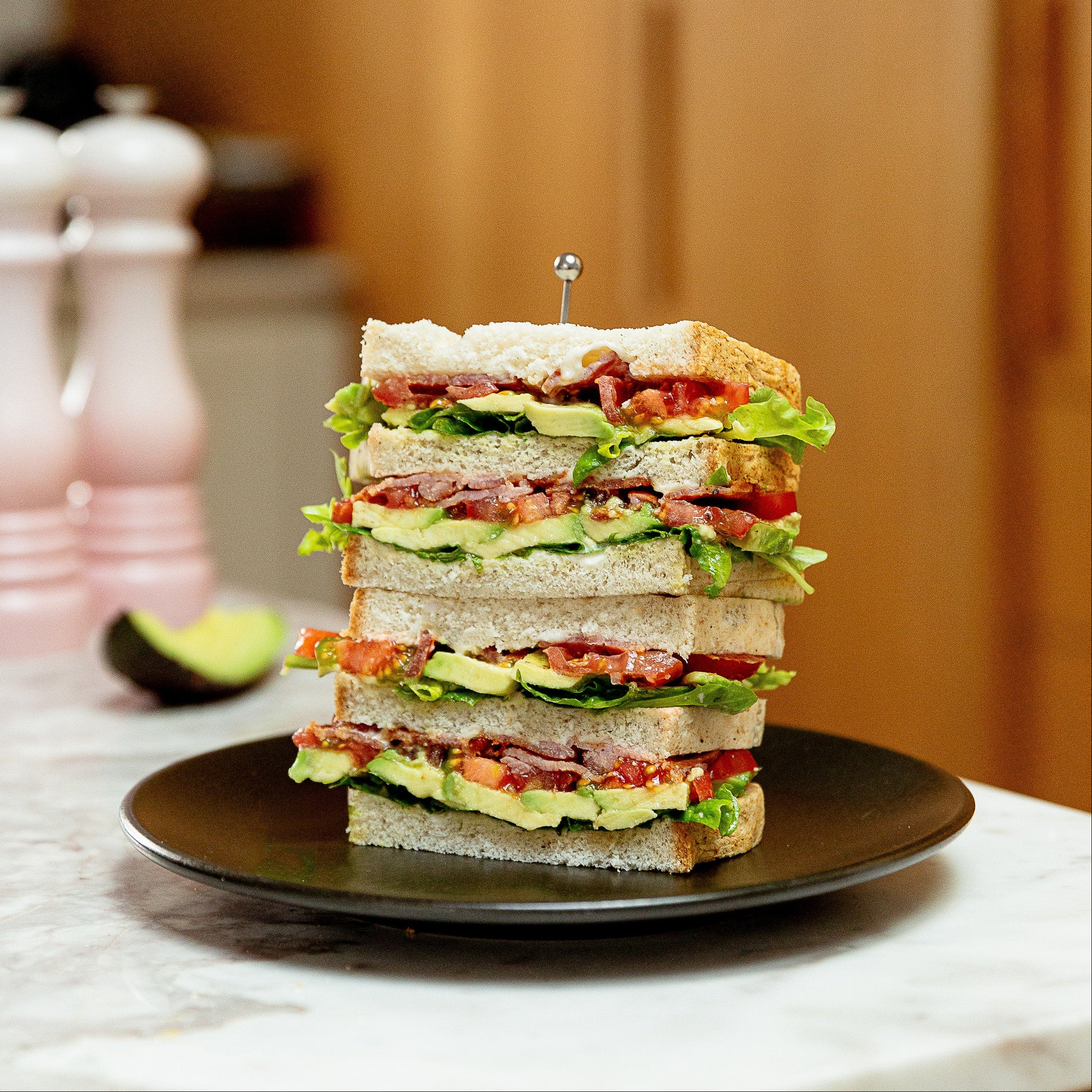 A tall stack of four club sandwiches on a black plate, set on a marble countertop with a warm wooden kitchen background.