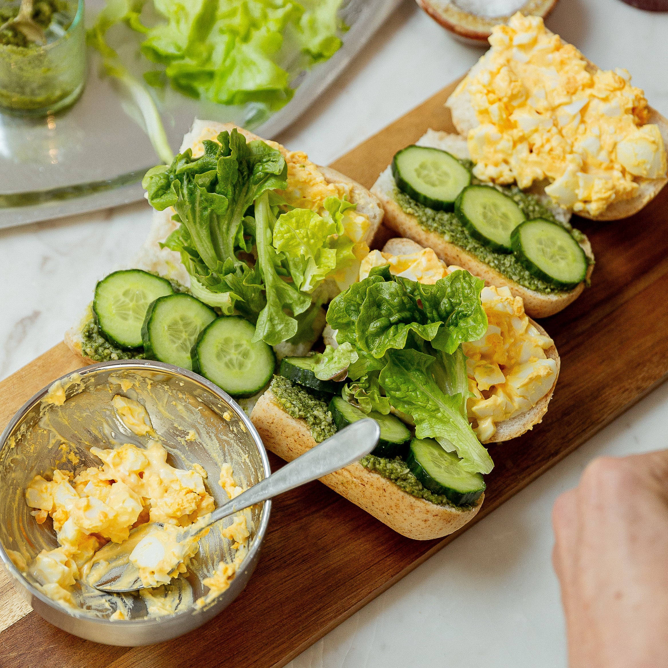 Open-faced sandwich bread topped with creamy yellow egg salad, green pesto, and sliced cucumbers, arranged on a wooden board with a small bowl of egg salad and fresh lettuce in the background.
