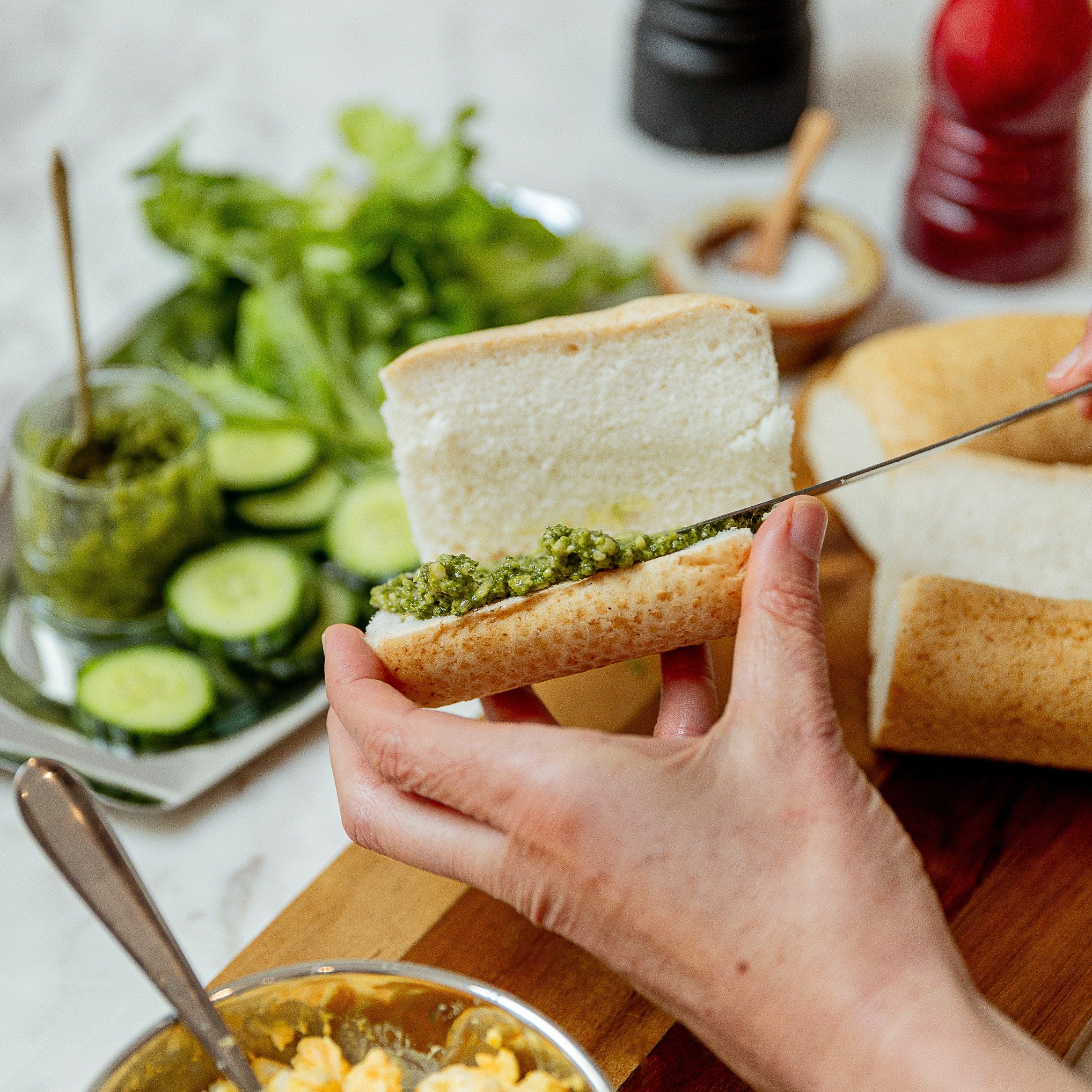 A close-up lifestyle shot of a person spreading green pesto onto a sliced white bread roll, with a bowl of egg salad, fresh cucumber slices, and greens in the background on a kitchen counter.