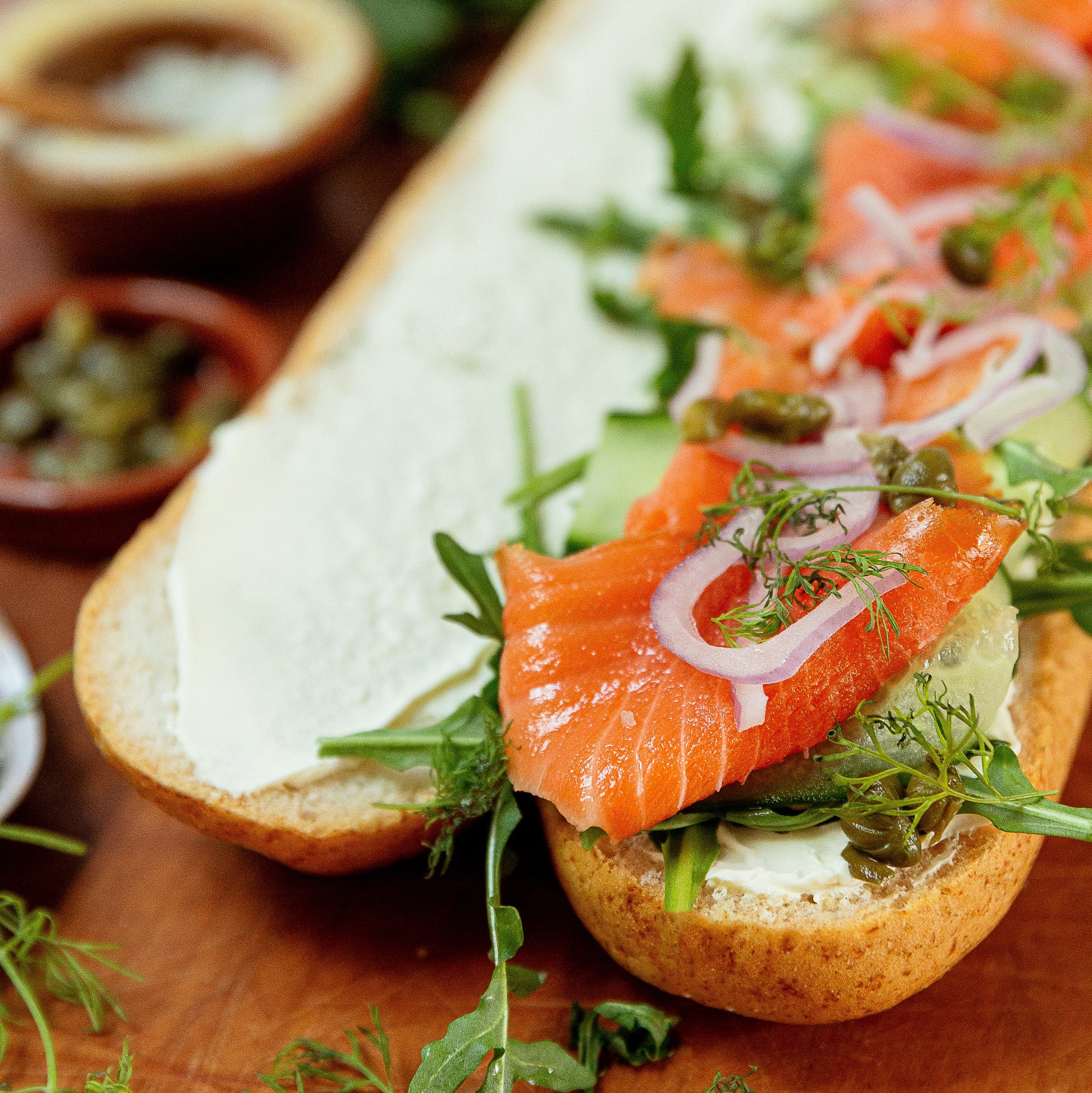 A close-up of a long sub sandwich on a wooden board, filled with salmon, arugula, red onion, capers, dill, and a thick white spread, with small bowls of seasoning in the blurred background.