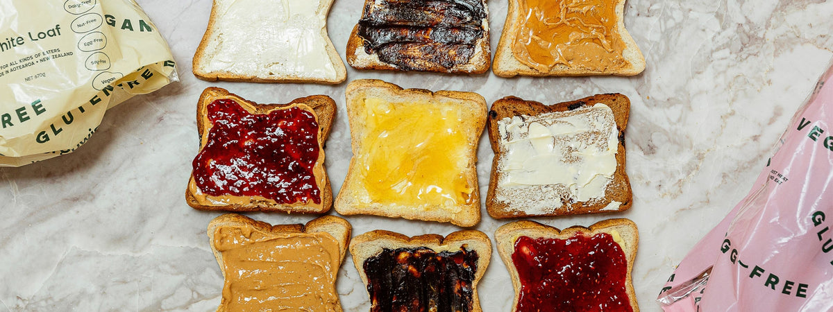 Nine slices of gluten-free white bread spread with various toppings including peanut butter, jam, and melted butter, arranged on a light marble surface with two partially visible bags of gluten-free bread at the edges.
