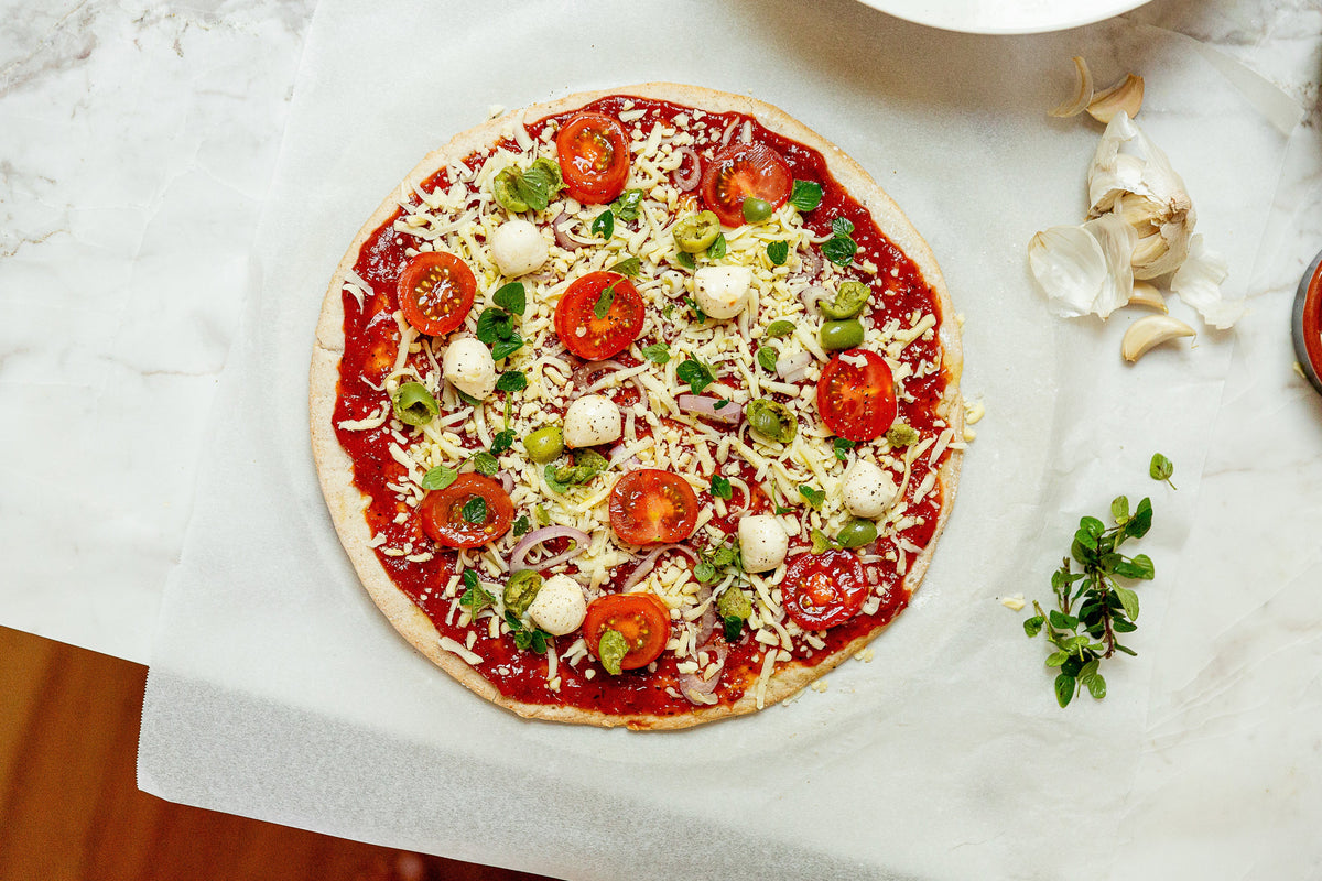 A pizza base with red sauce, cheese. tomatoes, fresh herbs and garlic on a marble bench.