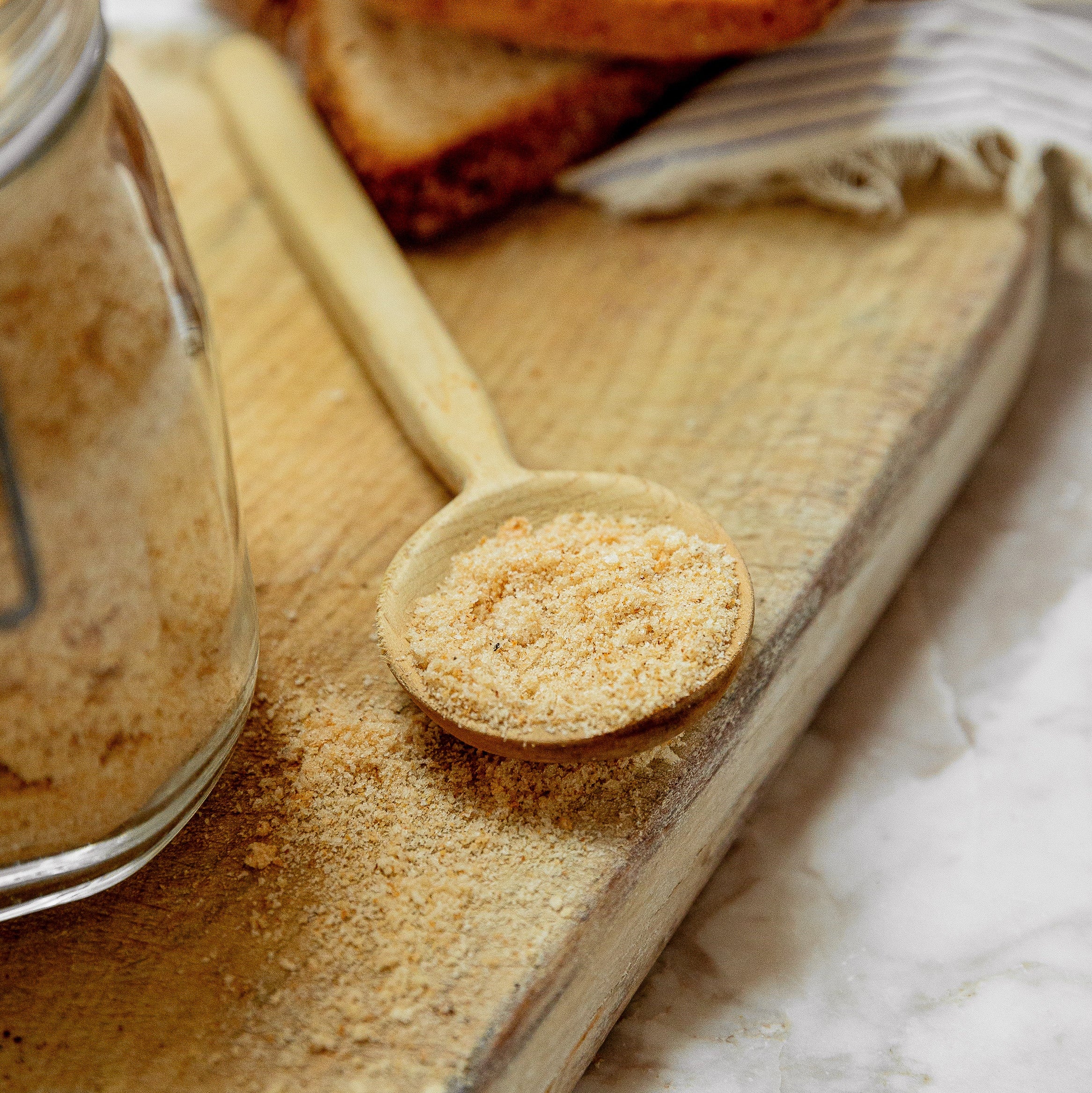 A close-up styled product shot of a clear glass jar filled with bread crumbs, with a wooden spoon holding more crumbs on a wooden board and sliced bread in the background.