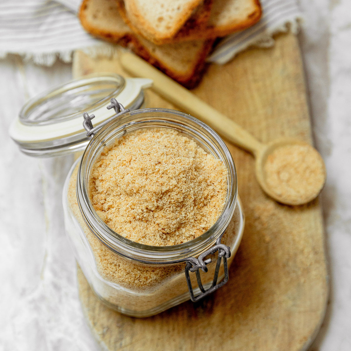 A clear glass jar filled with bread crumbs sits on a wooden board, with sliced bread and a small wooden spoon in the background on a white marble surface.