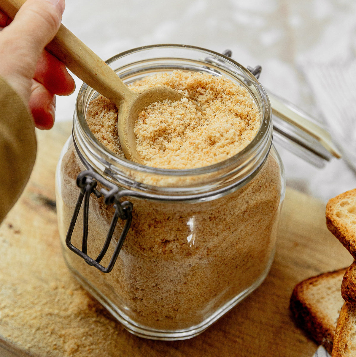A clear glass jar filled with bread crumbs shown open with a wooden spoon on a wooden board, with a hand holding the spoon and slices of bread nearby.