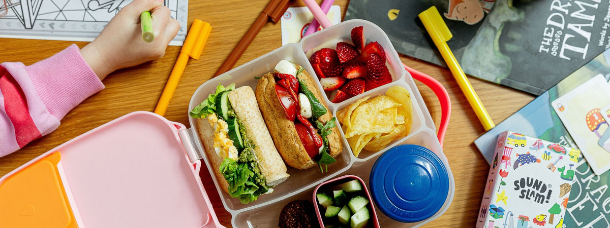 A colourful children's lunch box filled with bread rolls, strawberries, chips, cucumber salad, and a blue lid, shown on a wooden desk with books, crayons, and a child's hand holding a green crayon.