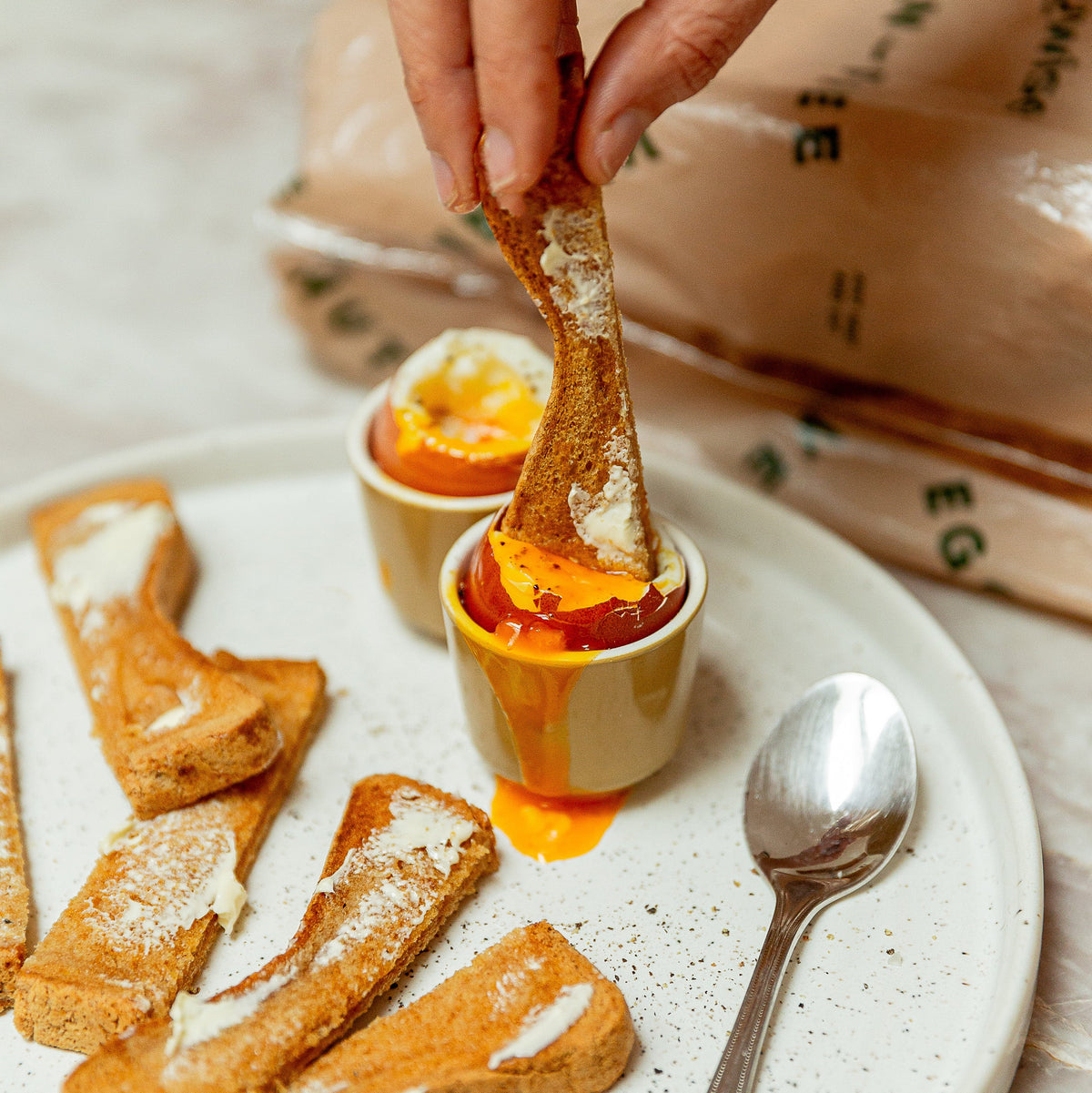 A close-up of a hand dipping a piece of toasted bread into a small cup of soft-boiled egg with orange yolk, served on a white plate with more toast and a spoon. A brown bag is blurred in the background.
