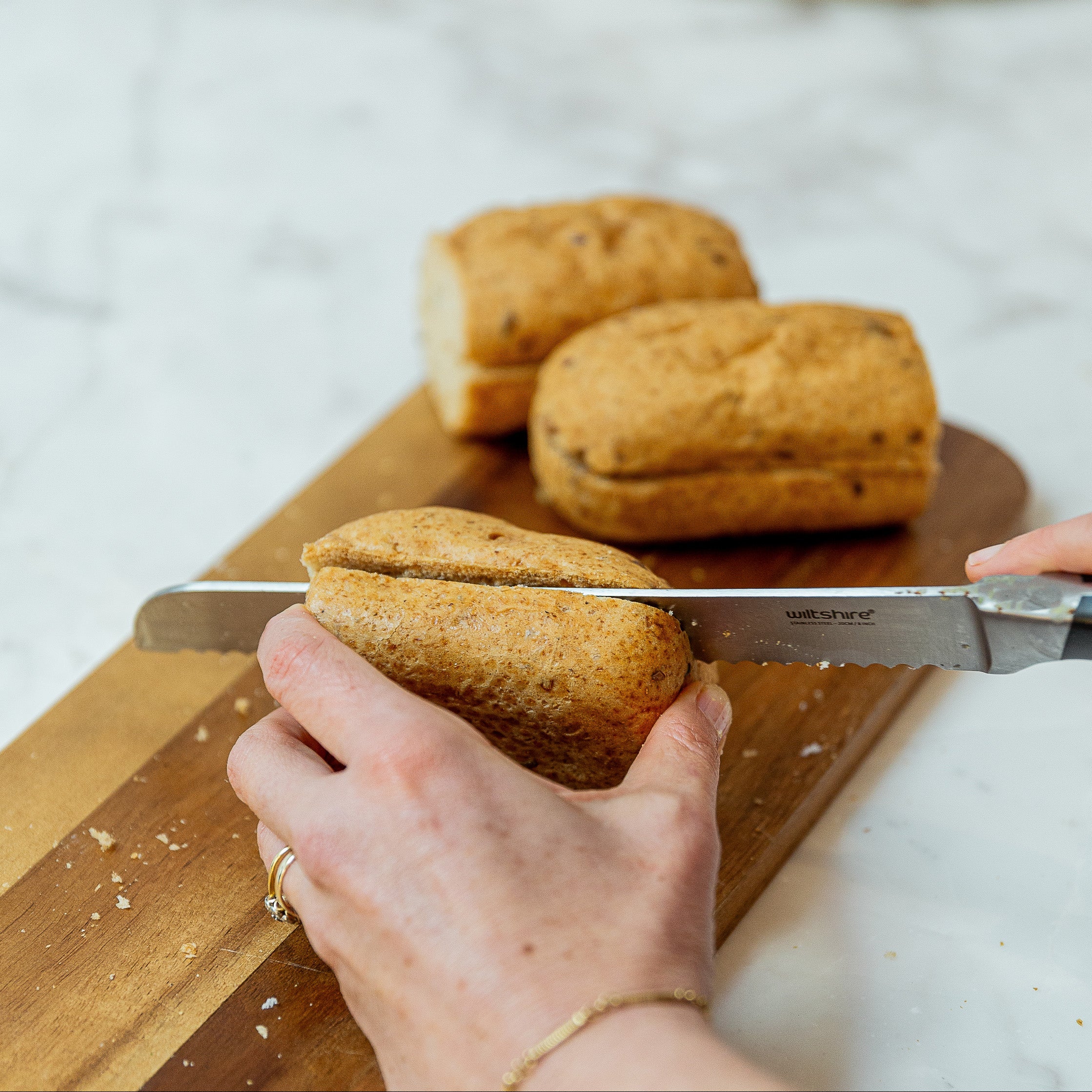 A close-up lifestyle shot of a person slicing brown bread roll on a wooden cutting board with a knife.
