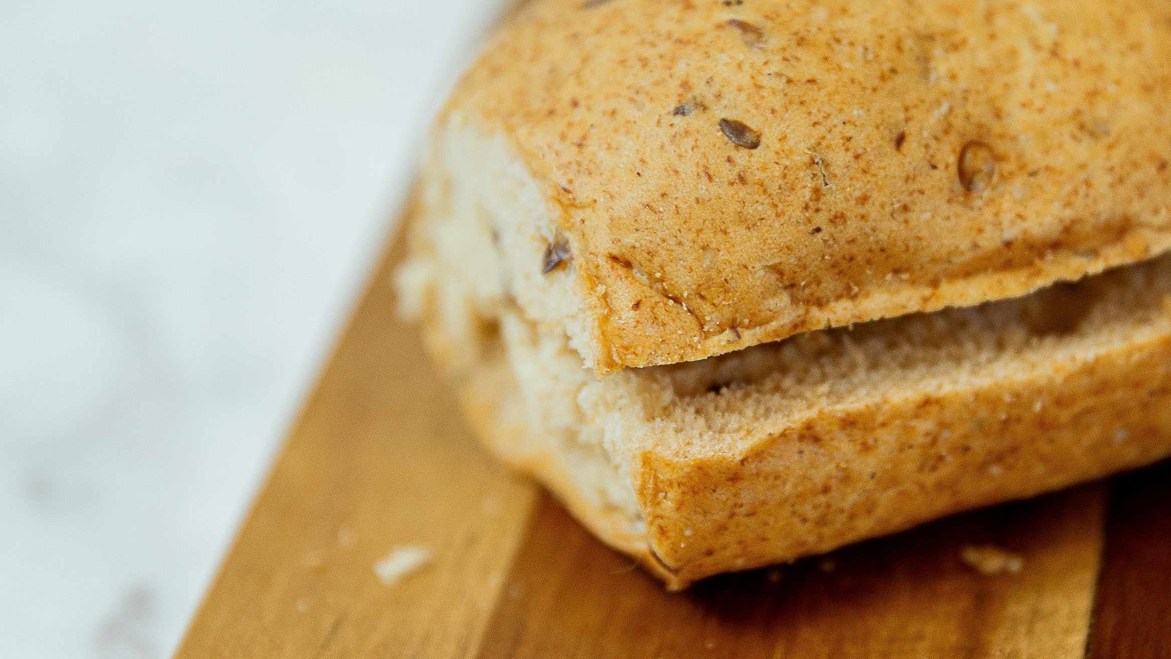 A close-up of a sliced bread roll with a golden-brown crust and visible dark seeds, resting on a wooden board with more rolls blurred in the background.