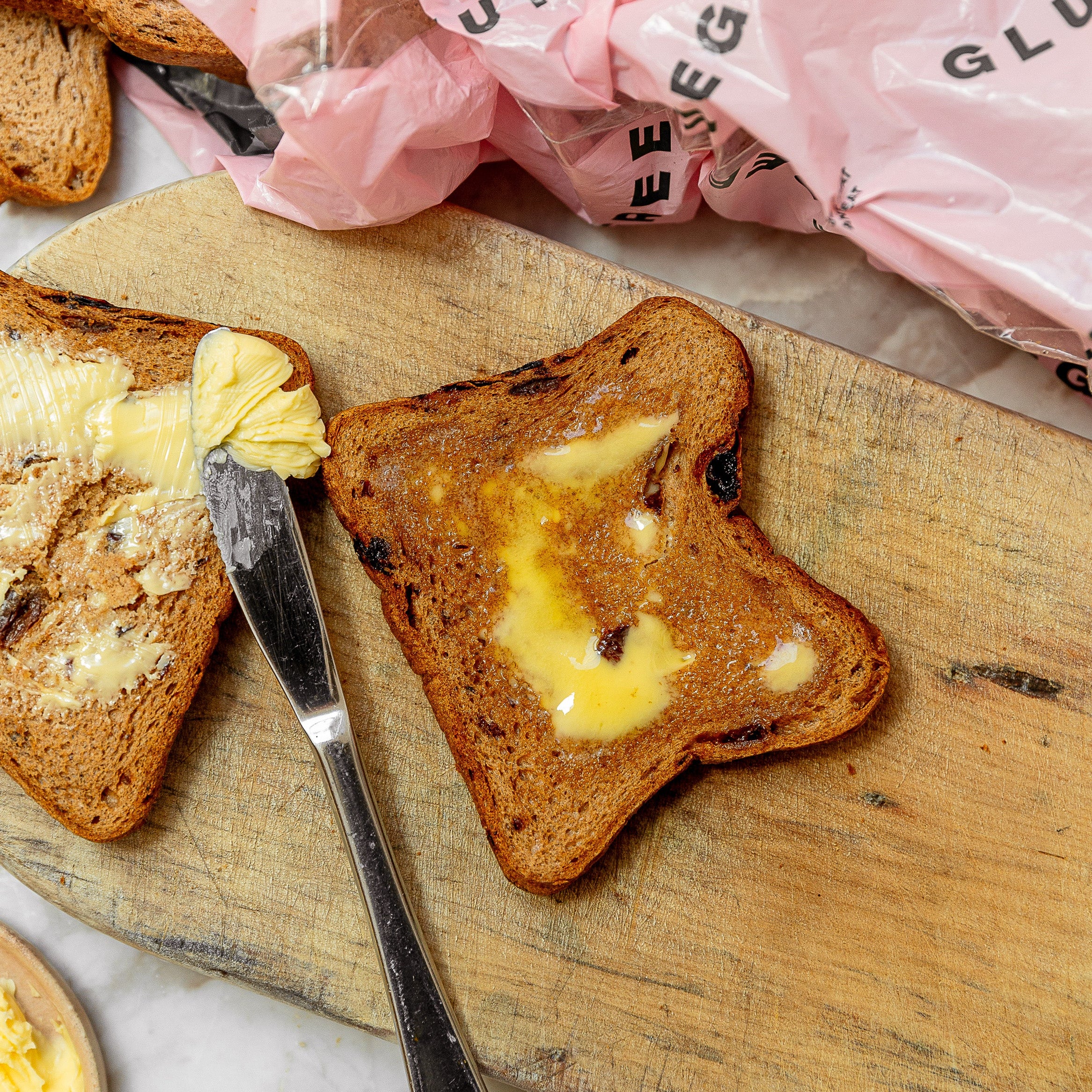 Two slices of toasted fruit bread on a wooden board, with butter and the other with a pink bread bag in the background.