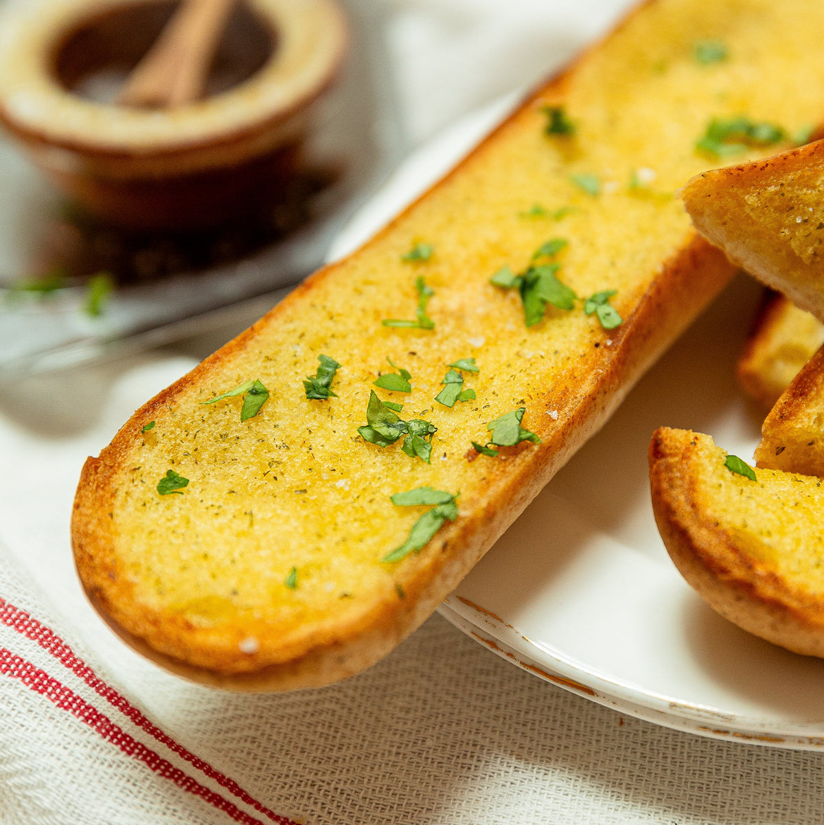 Garlic bread on a white plate, topped with melted butter, herbs, and coarse salt, with a small bowl of salt in the blurred background.