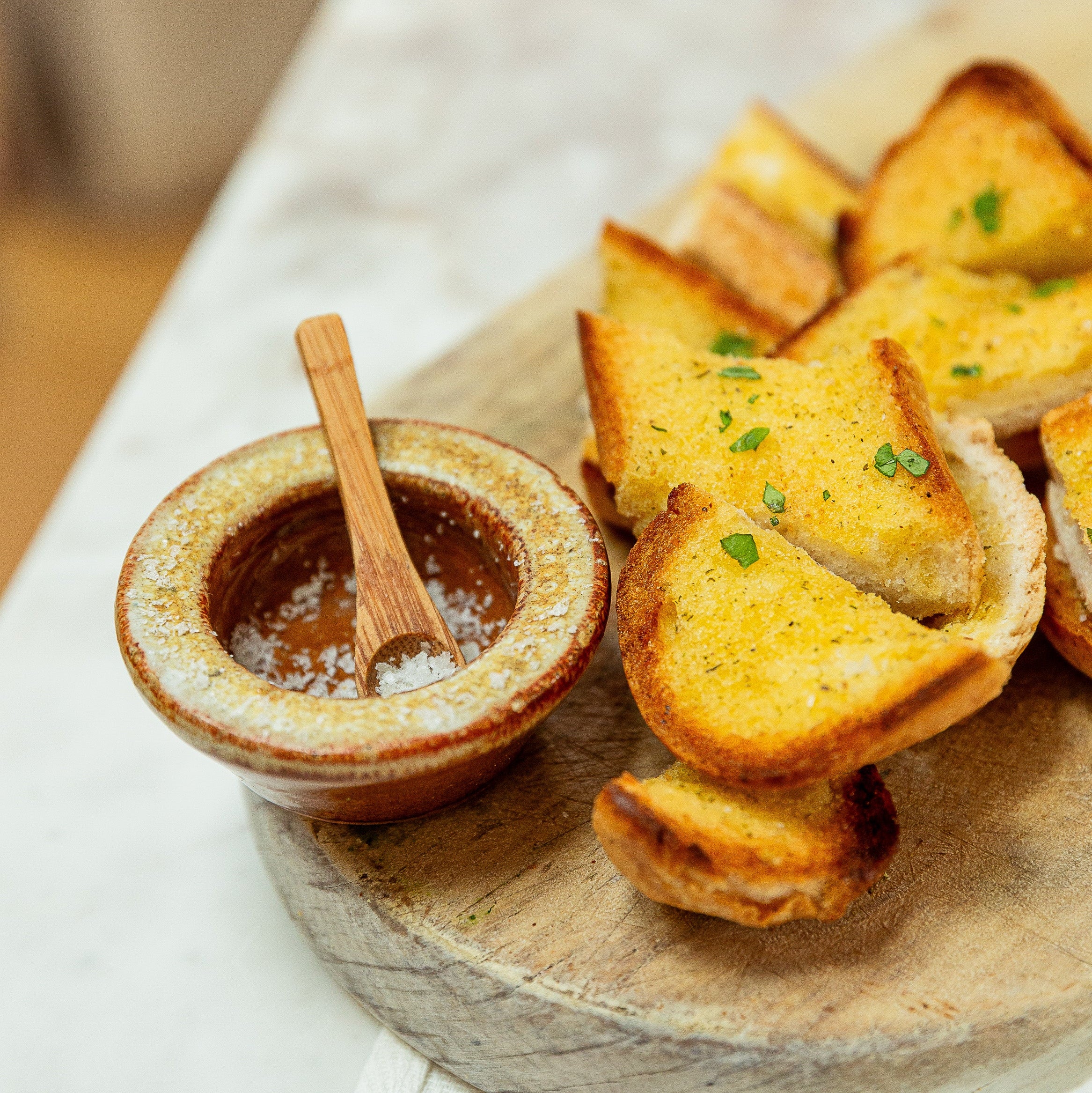 Toasted garlic bread slices on a round wooden board, served with a small ceramic bowl of salt and a wooden spoon, on a white tablecloth.