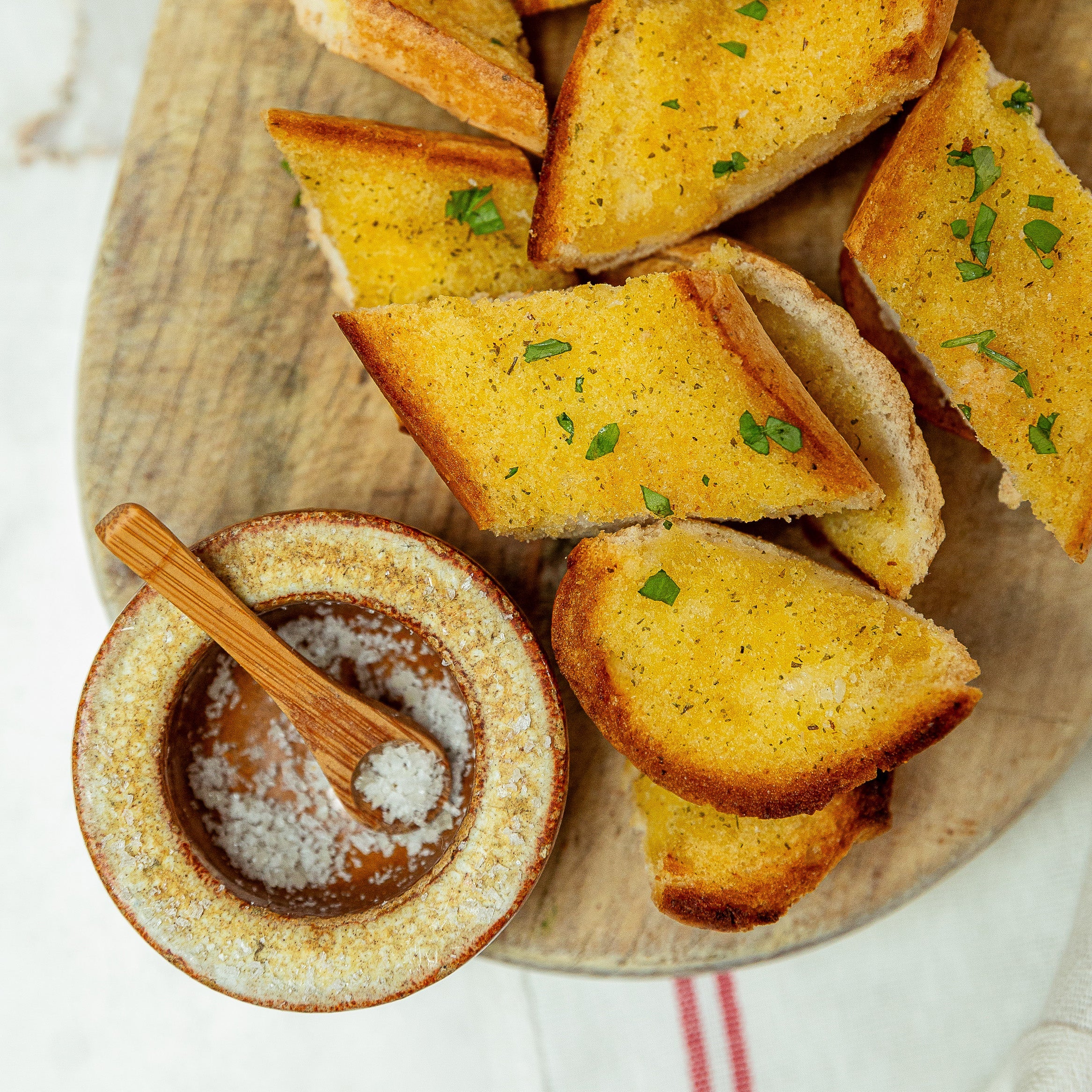 Sliced garlic bread on a wooden serving board with a small bowl of salt and a wooden spoon, garnished with chopped parsley.