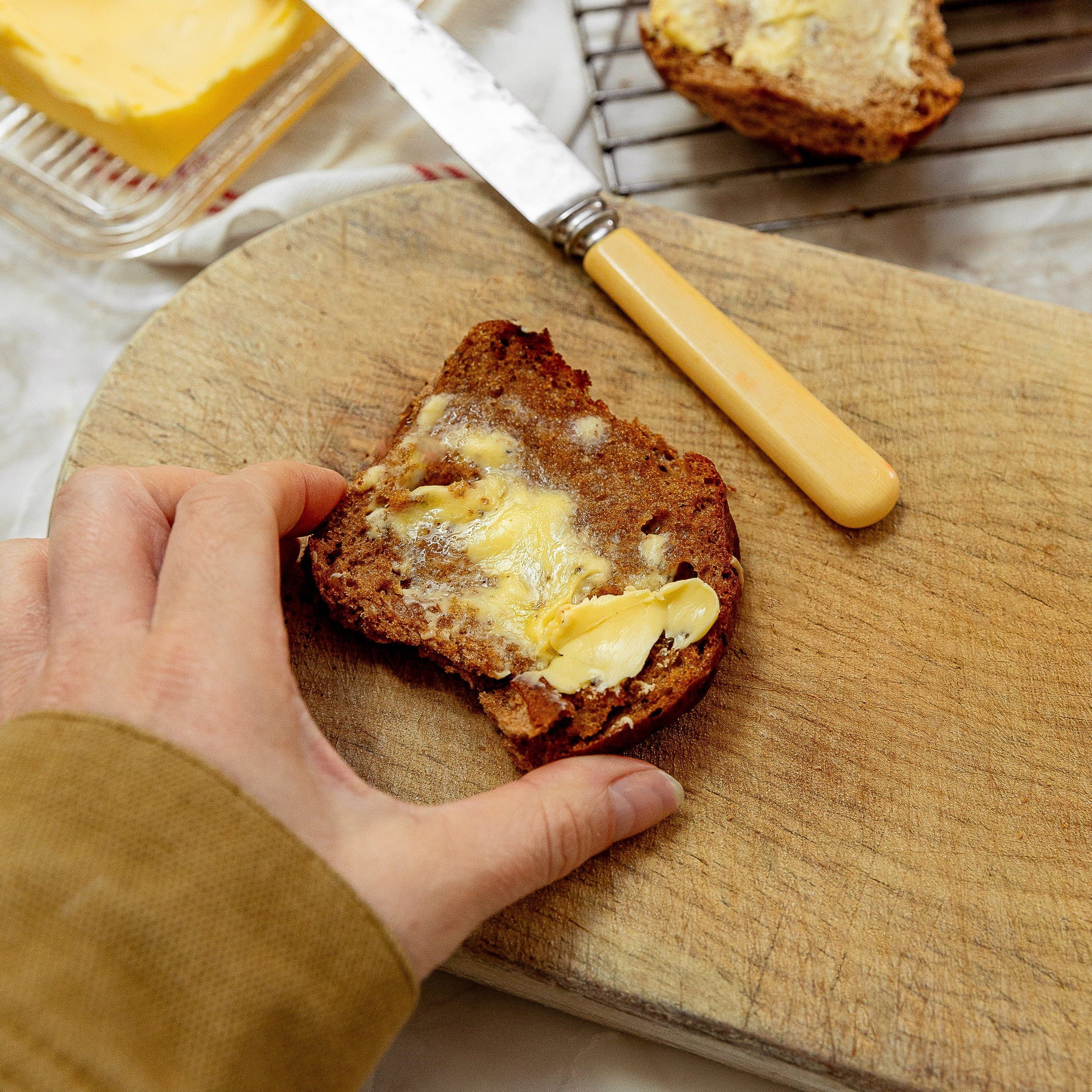 A hand spreads butter onto a slice of dark bread on a wooden cutting board, with a butter knife and more buttered hot cross bun in the background.