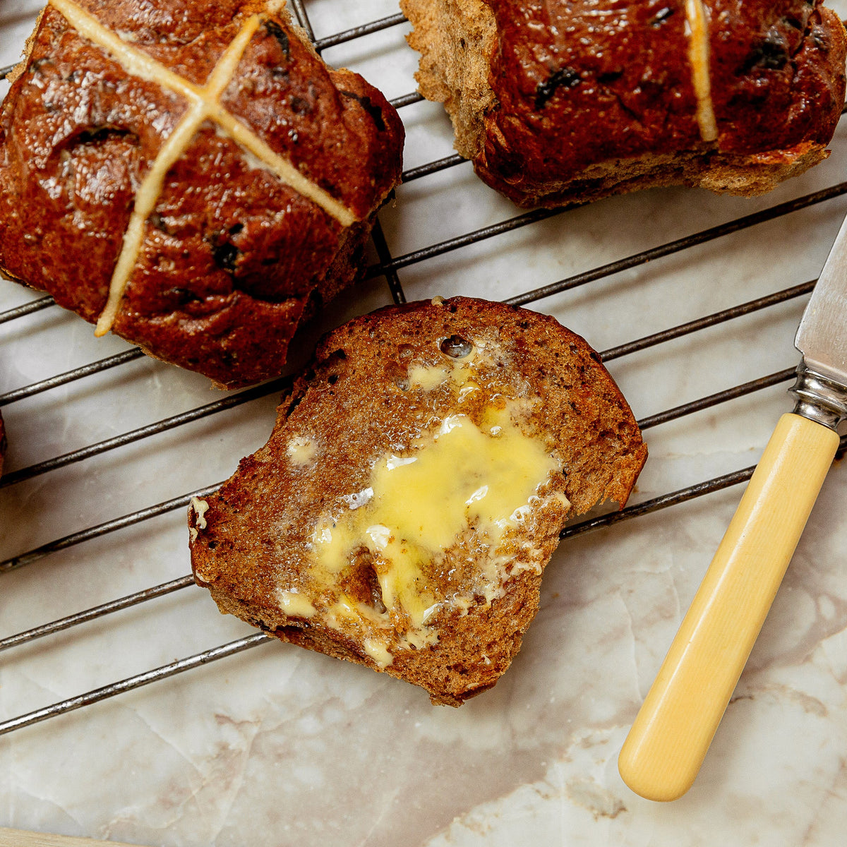 Close-up of several brown hot cross buns on a metal cooling rack, with one slice cut open and buttered. A butter knife with a pale yellow handle rests on a light marble-like surface.