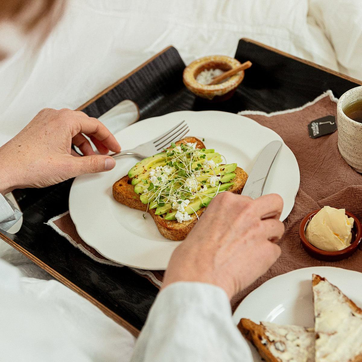 A breakfast scene in bed with a person serving avocado toast on a black tray. Plates, a mug, and additional toast are arranged on a dark tray in a softly lit bedroom setting.