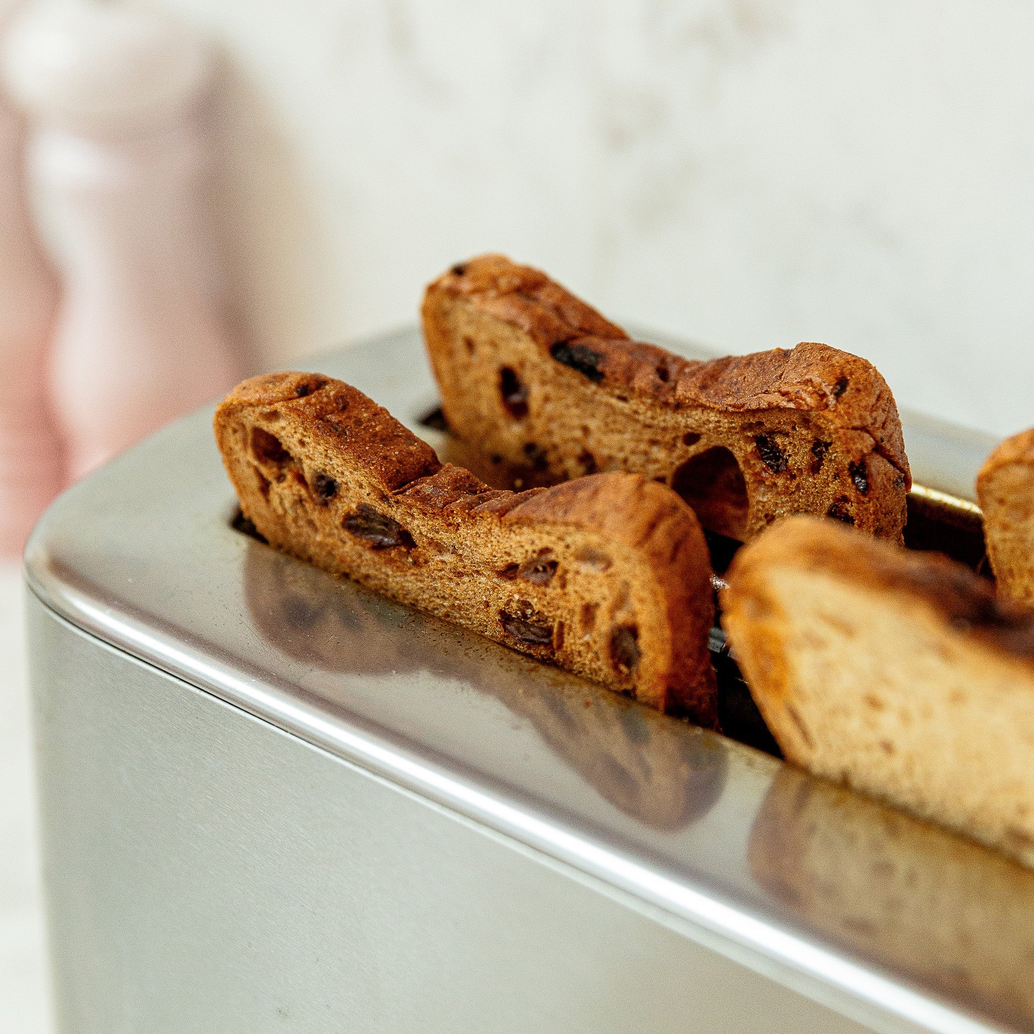 A close-up of a stainless steel toaster with several slices of fruit toast emerging from the slots, set against a softly blurred light background with pink salt and pepper shakers out of focus.