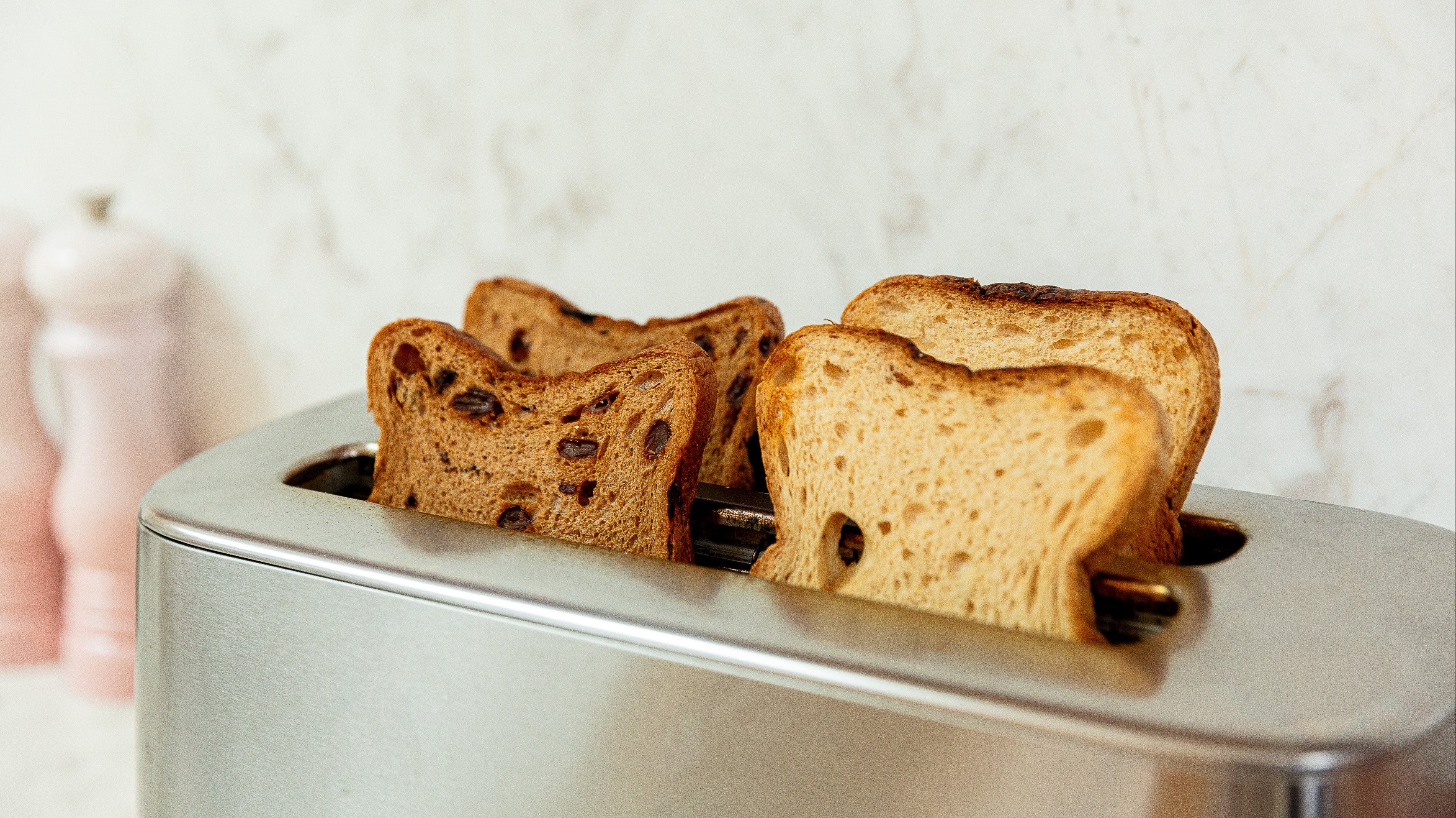 A stainless steel two-slot toaster with four slices of toasted bread with dark fruit pieces, set against a light marble-patterned background.