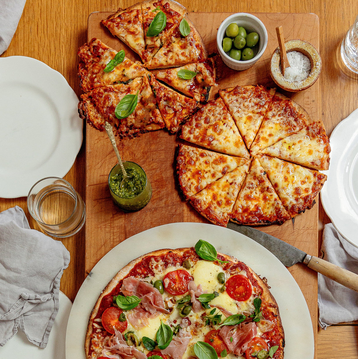 Three freshly baked pizzas arranged on a wooden table with plates, glasses, and small garnish bowls. One large pizza in the foreground has tomato, ham, cheese, and basil, while two smaller pizzas sit on a cutting board behind it with toppings like green olives and pesto.