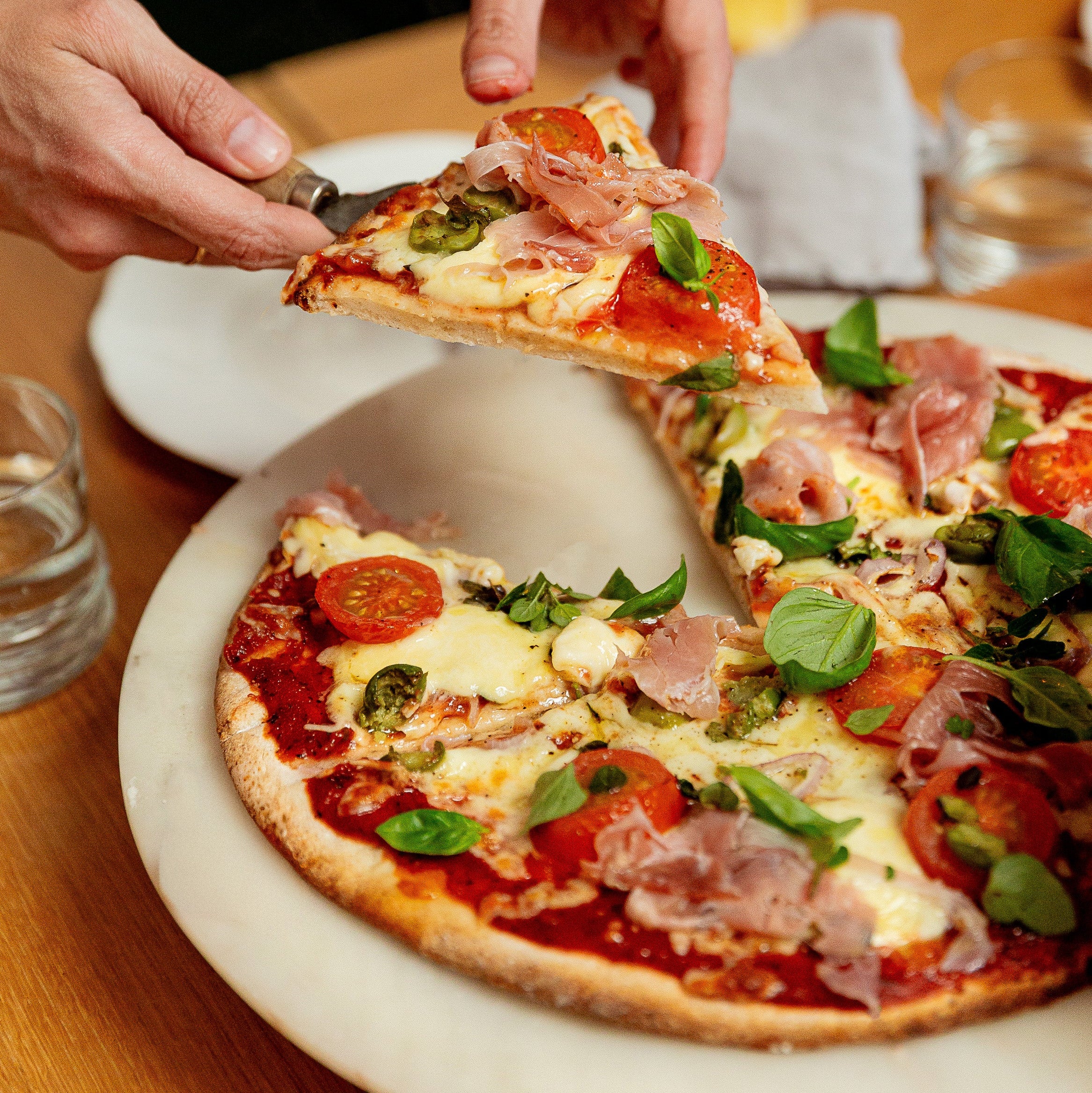 A close-up of a pizza on a white plate at a wooden table, with a slice being lifted by hands using a pizza cutter. The pizza is topped with tomato sauce, melted cheese, prosciutto, green basil leaves, and pepperoni.