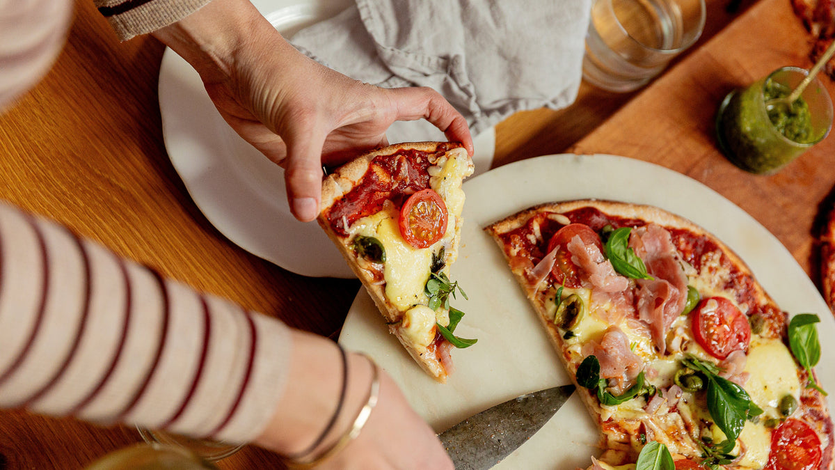 A close-up lifestyle shot of a person holding a slice of pizza with tomato, mozzarella, and basil on a wooden dining table next to a larger pizza on a stone plate.