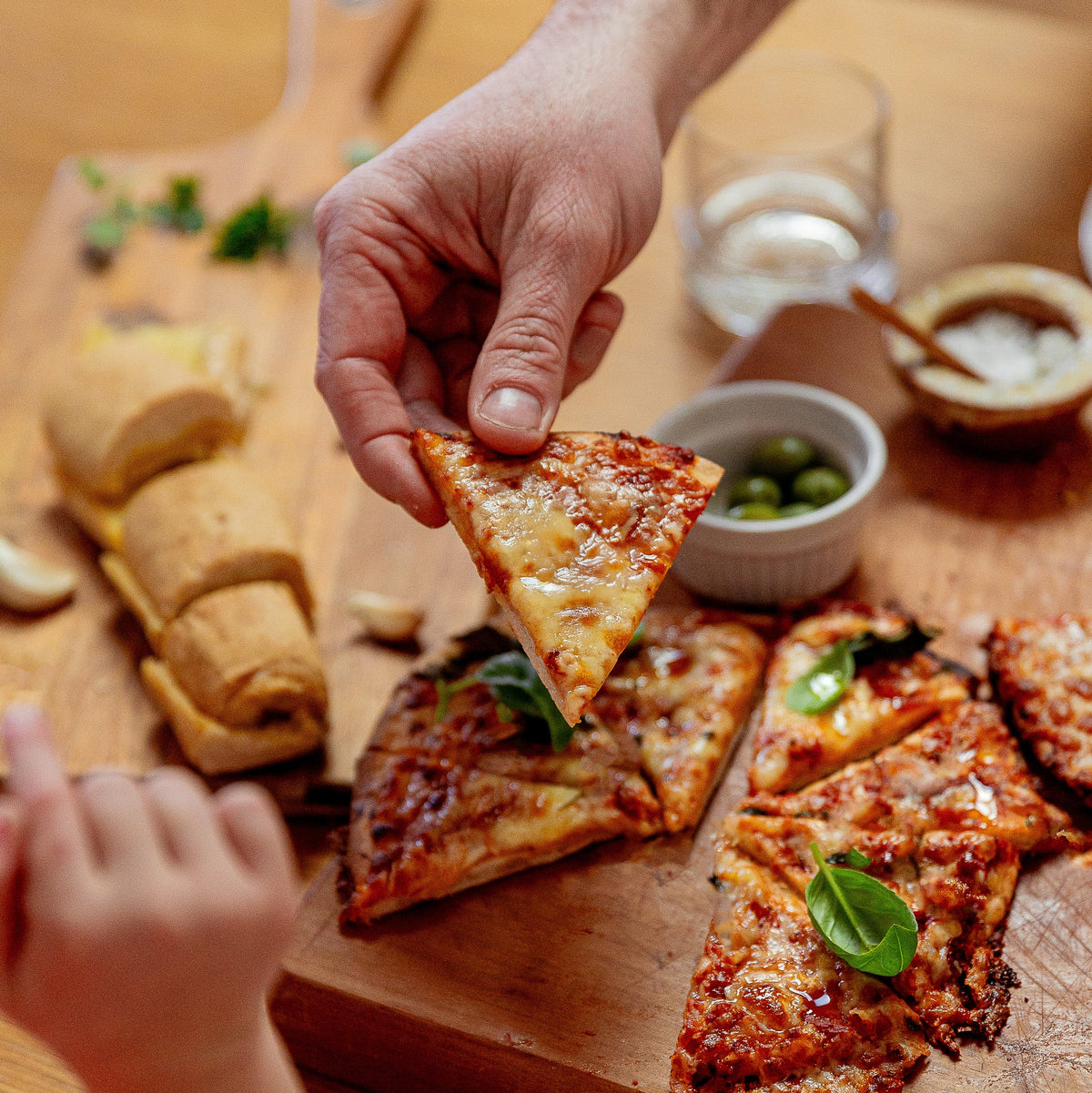 A close-up lifestyle shot of a hand holding a slice of cheesy pizza above a wooden board filled with pizza slices, bread, olives, and dipping sauce on a wooden table.