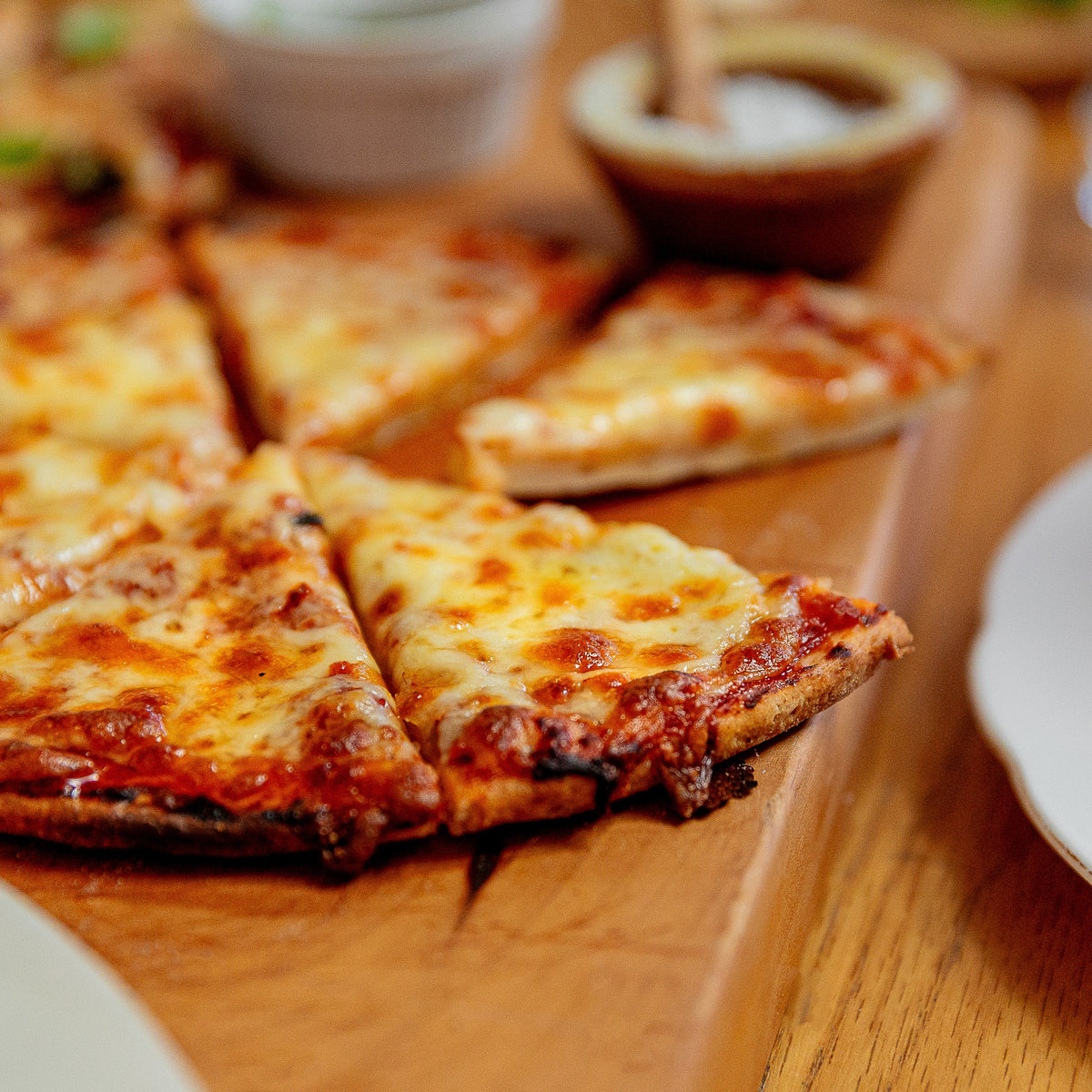 A close-up of a freshly baked cheese pizza on a wooden board, with several slices cut and one slice leaning against the crust. Small dipping sauce bowls and white plates are visible on a wooden table.