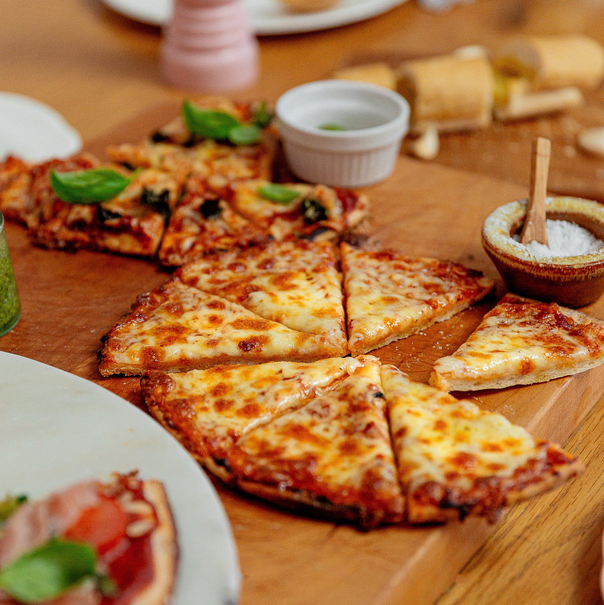 Cheesy pizza slices arranged on a wooden board on a dining table, with a few pieces topped with fresh basil leaves and small dipping dishes nearby.