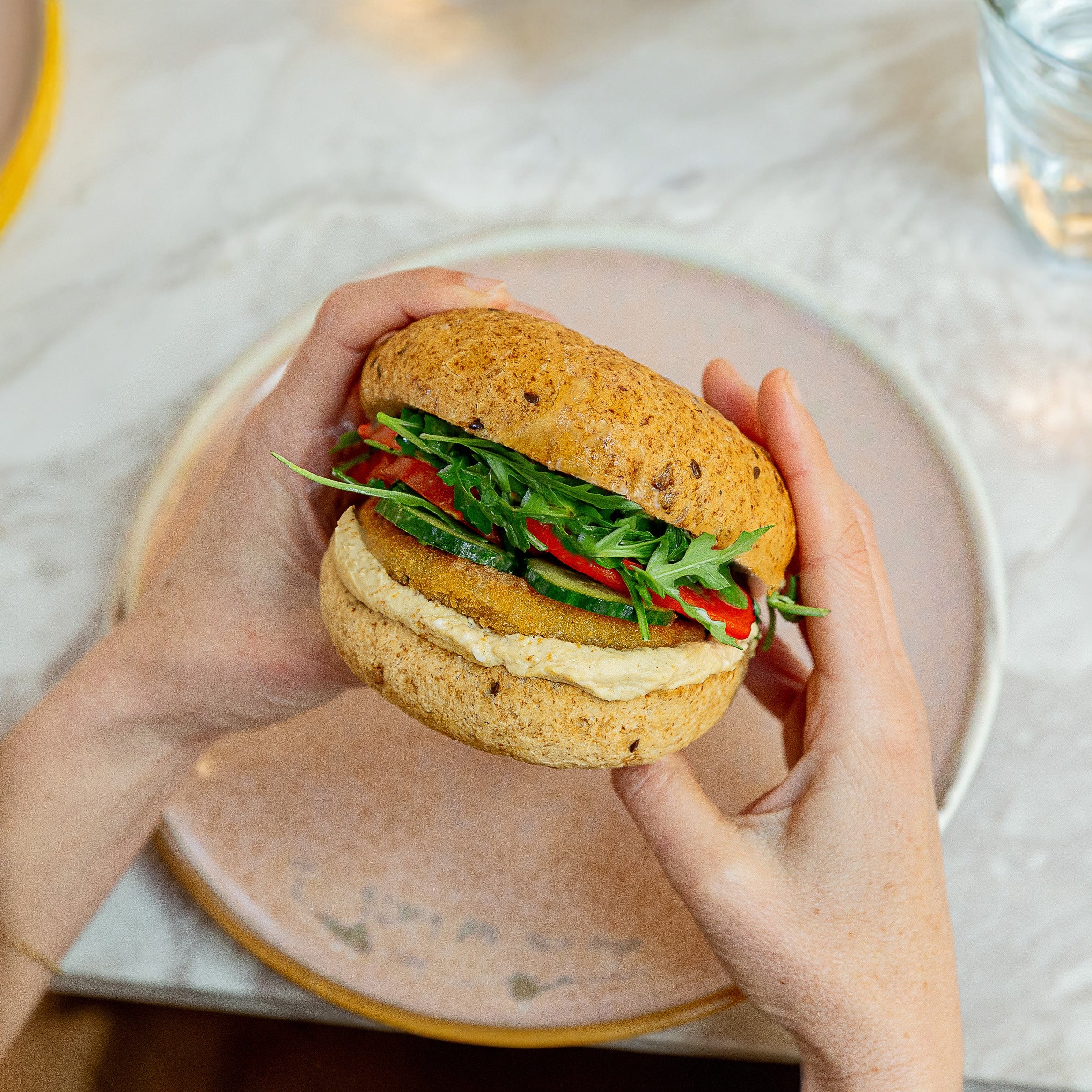 A person holds a gourmet sandwich with a thick veggie patty, fresh green arugula, red peppers, and a creamy sauce between two buns over a pink plate on a marble table.
