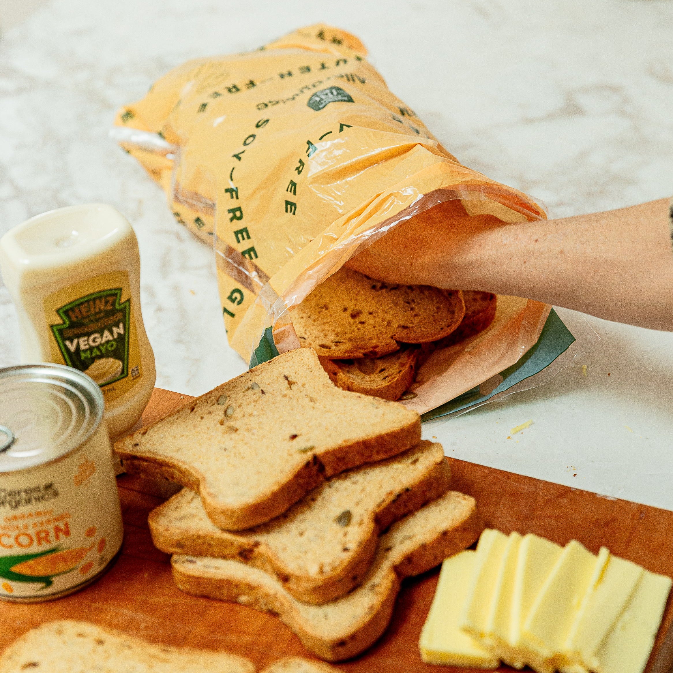 A person's arm and hand are shown unpacking vegan seeded bread from a orange Allergywise bag on a marble countertop, with slices of bread, butter, and a can of vegan mayo visible.